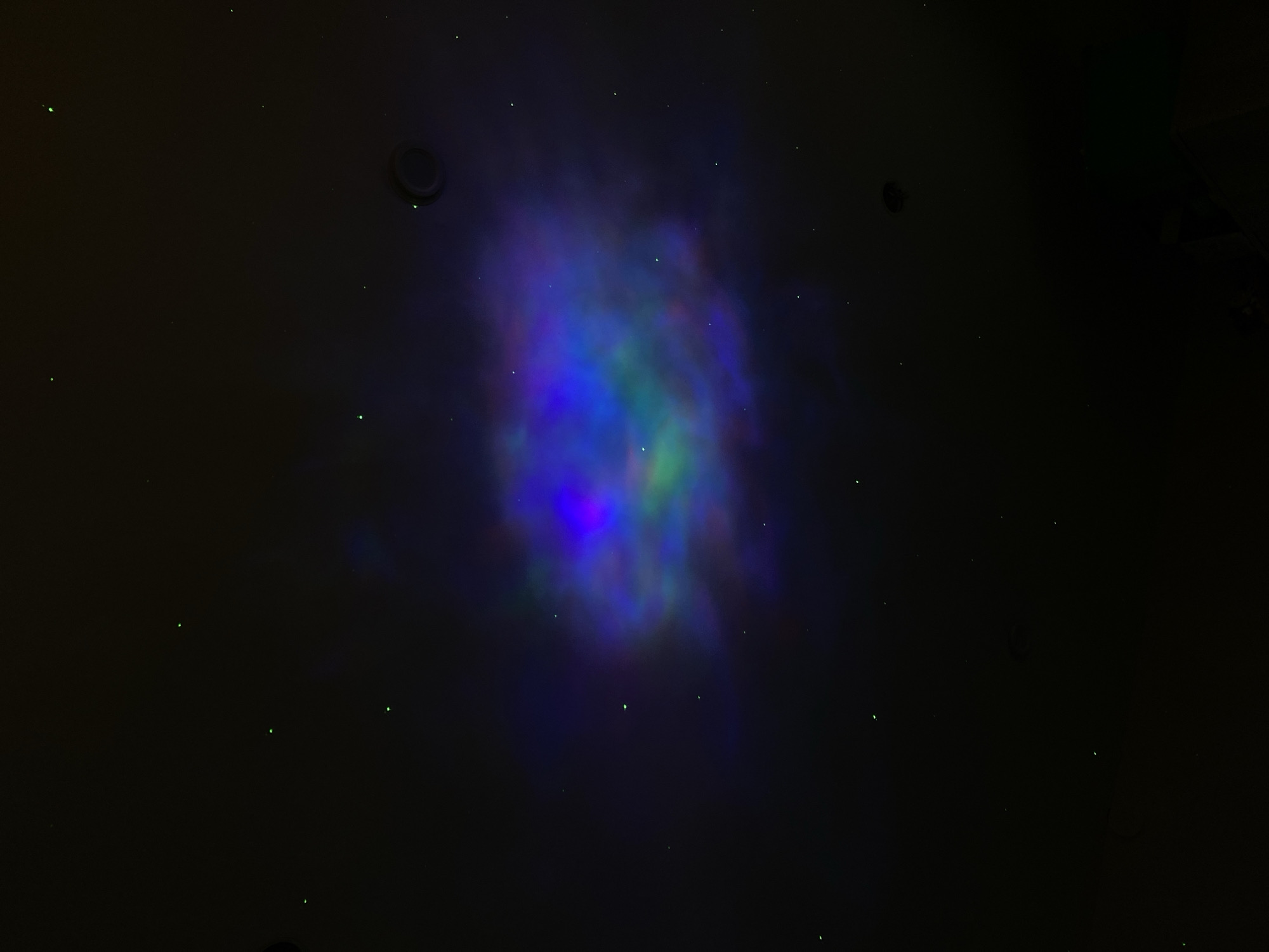 The image shows a dark, likely indoor, space with a nebula-like projection of blue, green, and purple light in the center. There are also small green dot-like lights scattered across the ceiling. A circular object, possibly a light fixture or sensor, is visible on the ceiling to the upper left of the nebula projection. The right edge of the frame is mostly black and obscured. There is no information in the image to determine the location in Votkinsk, Russia, or any details about people, subjects, or actions.