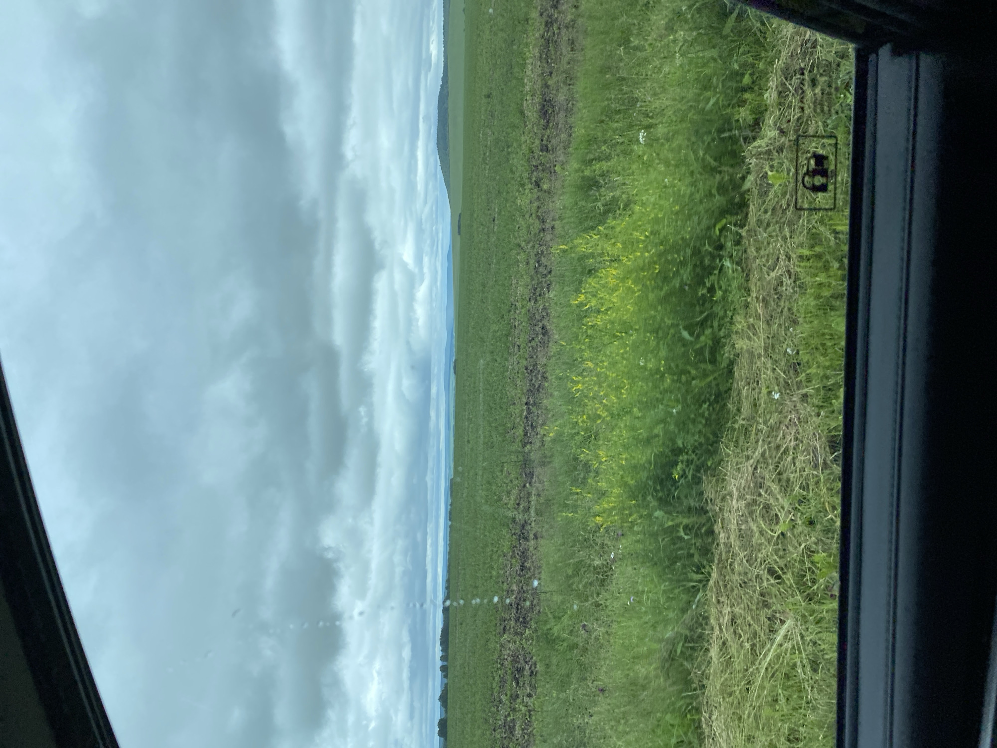 The image depicts a cloudy sky over a rural landscape. In the foreground, the edge of a car window is visible, with a car door's interior panel at the bottom. Through the window, a field of green grass and vegetation stretches into the distance, with a section of tilled earth in the middle ground. Beyond the field, a tree line and rolling hills can be seen under a dramatic, overcast sky. A small symbol, appearing to be a child lock icon, is present on the car window. The scene is likely from Kliuchiki, Russia, given the context.