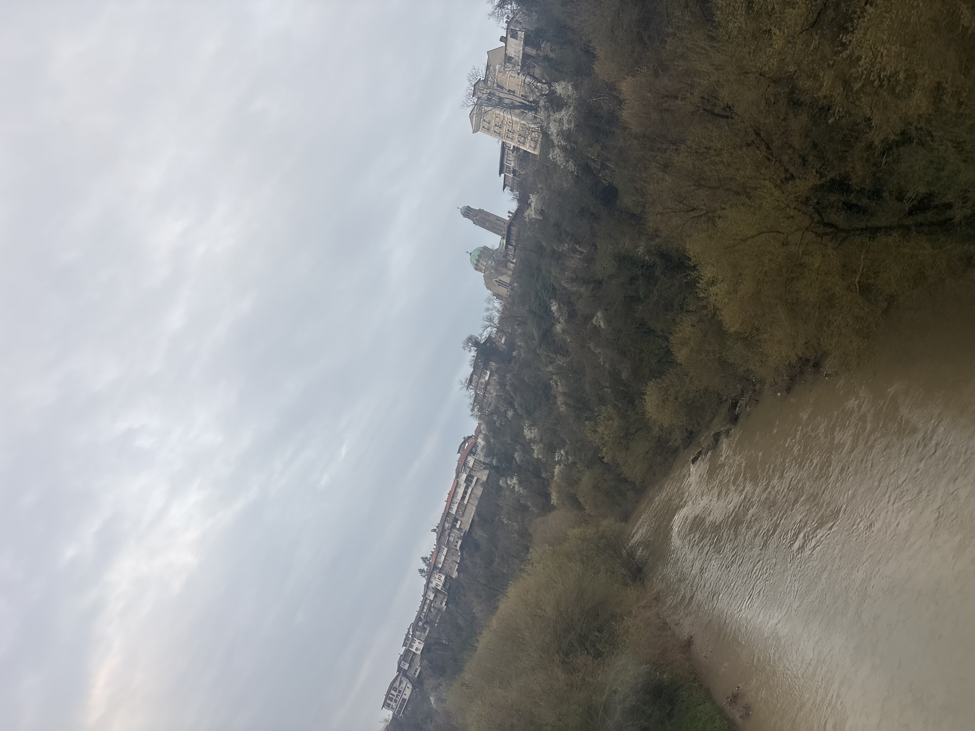 The image depicts a scene in Veliko Tarnovo, Bulgaria. The Yantra River flows in the foreground, its water appearing muddy. Along the river's edge is vegetation, primarily trees with early spring foliage. Rising steeply from the riverbank is a cliff on which a series of buildings are perched. These buildings appear to be houses and apartments, and further up the cliff is a church with a prominent bell tower and a green dome. The sky is overcast, suggesting a cloudy day. No people are visible in the scene.
