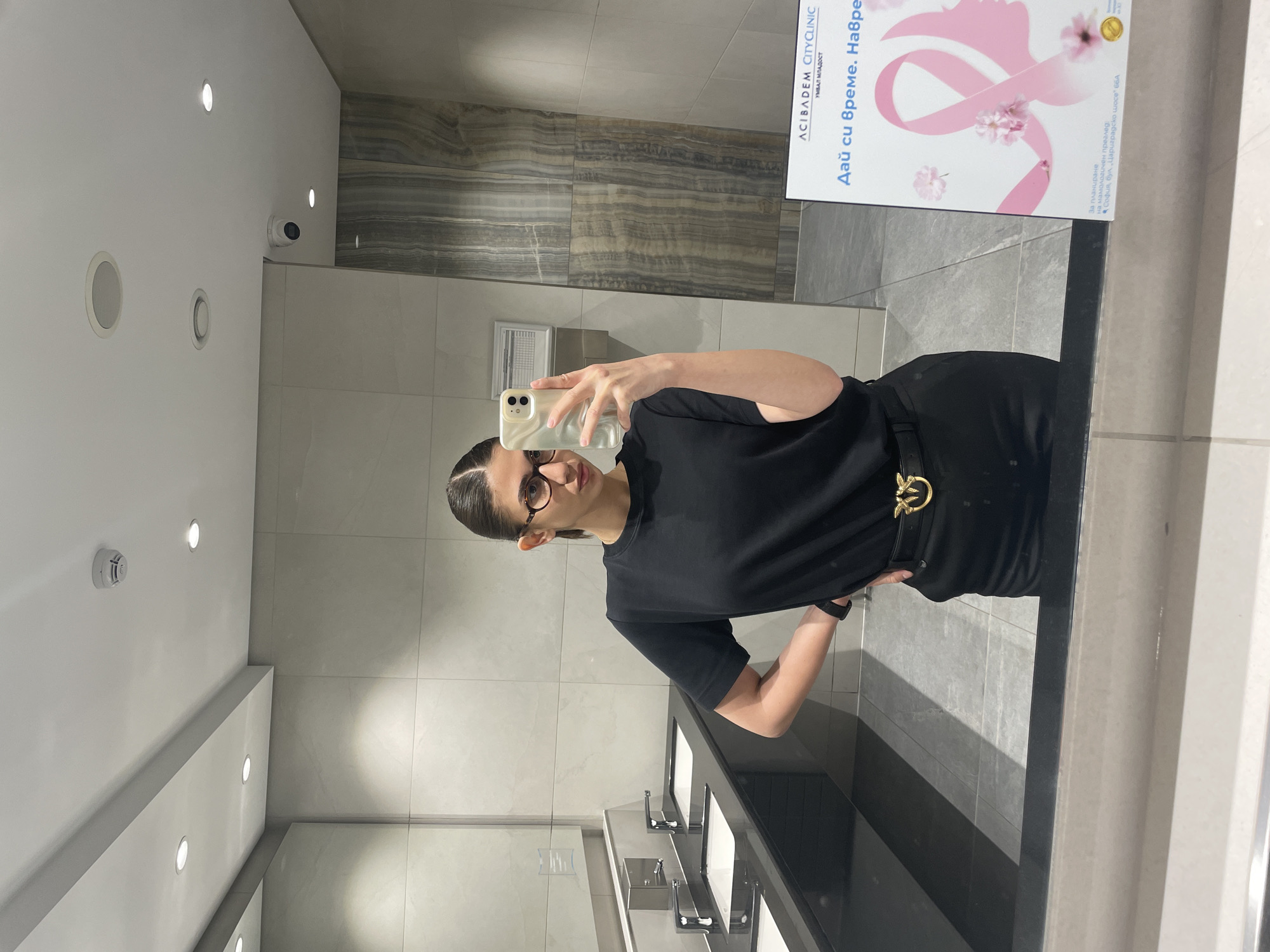 The image shows a woman taking a selfie in a bathroom. The bathroom appears to be in a public place, as there are multiple sinks visible. There is a sign on the wall for ACIBADEM CITYCLINIC, located in Sofia, Bulgaria. The sign is advertising mammography services for breast cancer screening. The woman is wearing a black outfit. The environment includes tile walls, recessed lighting, and modern fixtures.