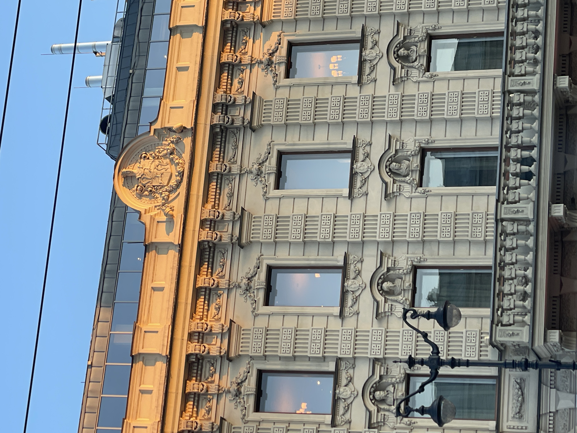 The image depicts the facade of a building in Saint Petersburg, Russia. The architectural style exhibits ornate detailing with windows framed by decorative elements, including sculpted figures. A modern glass addition abuts the older building. A street lamp is visible in the foreground. The sky is clear.