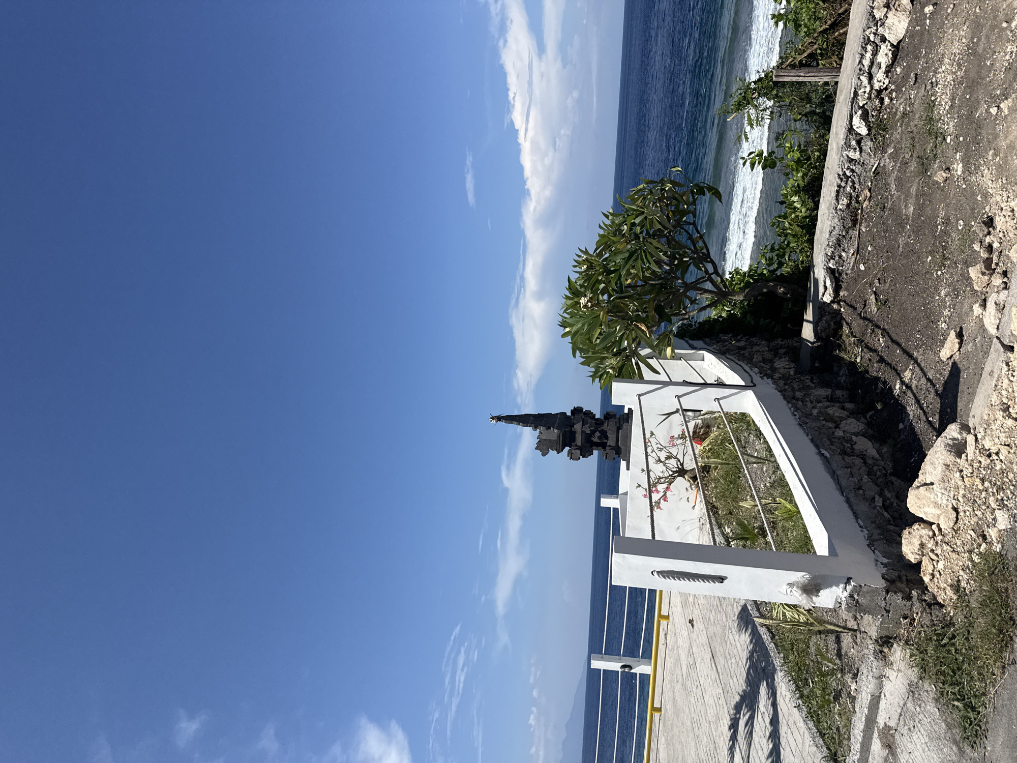 The image depicts a coastal scene in Batununggal, Indonesia. A white fence overlooks the ocean. A black stone monument is positioned on the other side of the fence. Vegetation, including a small tree, grows along the edge of the cliff. The sky is blue with scattered white clouds. The ocean is visible in the background. The scene appears to be a viewpoint or memorial site.