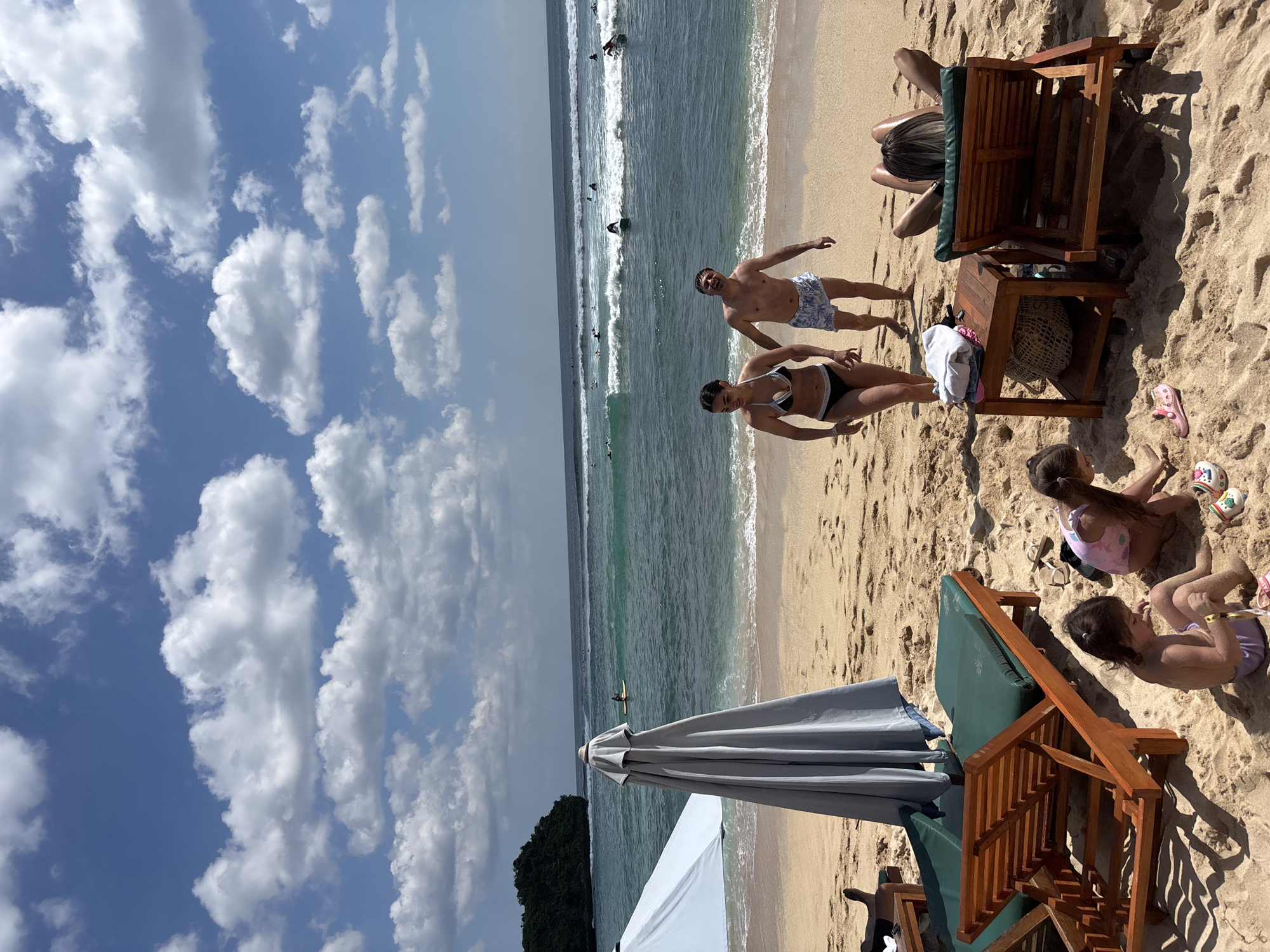 This image depicts a vibrant beach scene on a sunny day in Kuta Selatan, Indonesia. The upper portion of the frame features a bright blue sky dotted with numerous white cumulus clouds. Below the sky, the ocean transitions from a clear turquoise near the shore to a darker blue further out, characterized by multiple parallel waves breaking with white foam. Numerous dark figures, identifiable as surfers, are spread across the water, some actively riding waves on surfboards while others await them.
The foreground is dominated by light brown sand. On the left, a large, light blue beach umbrella is open, while a large, gray beach umbrella is folded and standing upright behind a wooden beach chair. Two wooden beach chairs with dark green cushions are present; one is partially visible on the far left, and another is seen from behind on the right, occupied by an individual lying down with their back to the camera. Between the right beach chair and the children, a small wooden side table holds folded light-colored clothing and a woven bag.
Two young children are seated directly on the sand in the foreground, engaged in play. One child wears a pink one-piece swimsuit, and the other wears a purple swimsuit bottom. Nearby, a pair of pink sandals and a pair of multicolored clogs are resting on the sand.
In the mid-ground, two adults are walking out of the ocean onto the sandy beach. The woman on the left wears a black and white bikini, and the man beside her wears blue and white patterned swim trunks. Both are barefoot and appear to be smiling or talking. On the far left, a lush, dark green, heavily vegetated landmass or small island juts into the ocean under the clear sky.