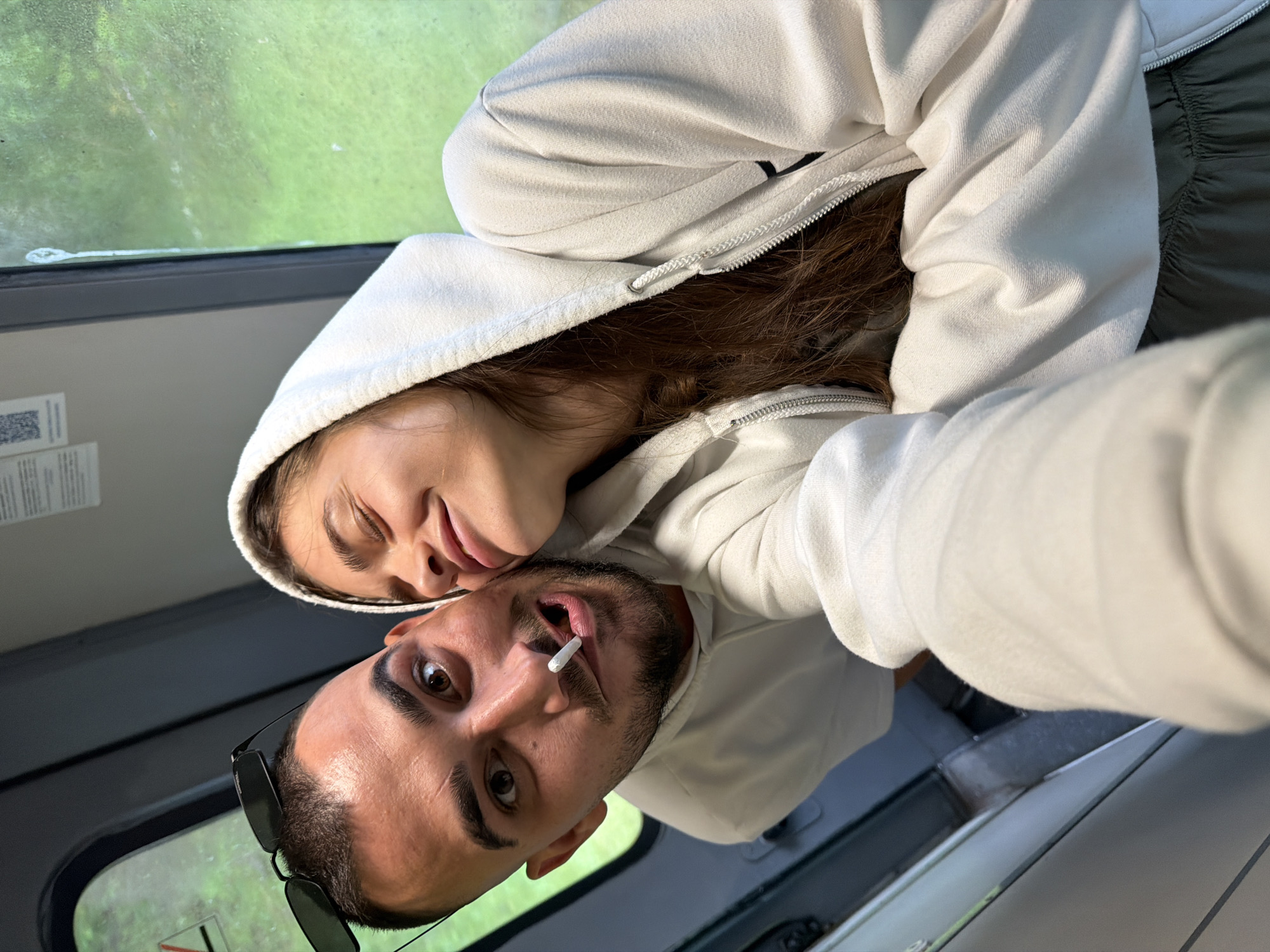 An overhead selfie captures a man and a woman in what appears to be the interior of a vehicle, possibly a car, bus, or train. The image is oriented sideways, but depicting the subjects correctly, the man is in the foreground, looking directly up at the camera. He has dark hair, a beard, and sunglasses pushed up onto his head. A rolled white cigarette is held in his mouth, slightly to the right.
Behind him, and leaning her head affectionately on his, is a woman with long brown hair. She is smiling softly with her eyes partially closed, wearing a white hooded sweatshirt, similar to the man's. Both individuals are dressed in light-colored hooded tops.
Through a window visible on the left side of the image (when oriented vertically), green foliage and blurred scenery suggest the vehicle is moving through a natural or semi-natural environment during daylight hours. A portion of a white surface with a black and white QR code or barcode is also visible near the window. The overall mood conveyed is one of casual intimacy and contentment, despite the cigarette in the man's mouth.