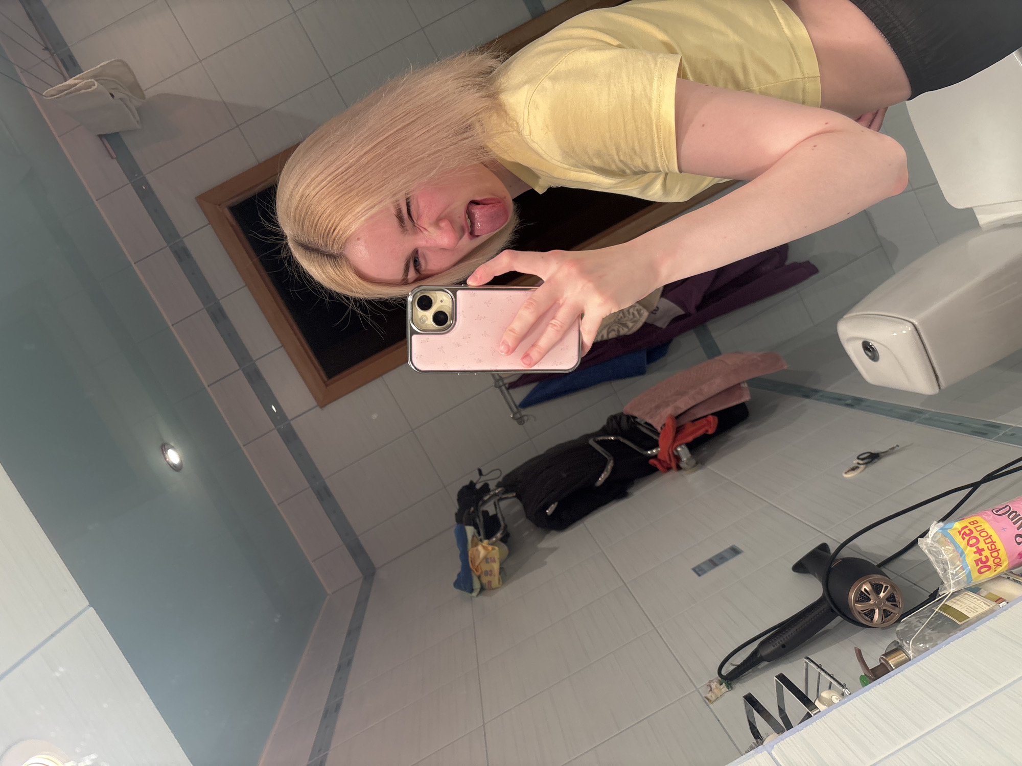 The media file is a selfie taken by a young woman in a bathroom. She has blonde hair and is wearing a yellow t-shirt and black shorts. She is sticking her tongue out and making a funny face at the camera. In the background, there is a toilet, a mirror reflecting the scene, and various bathroom items such as a hair dryer, scissors, and towels. The bathroom appears to be tiled with blue and white tiles. The lighting suggests it is daytime, though no windows are visible. The overall mood is playful and informal. The woman is holding a pink iPhone in her right hand to take the picture. Visible text includes "DANISH" and "BUOTRIBOK" on a package in the background.