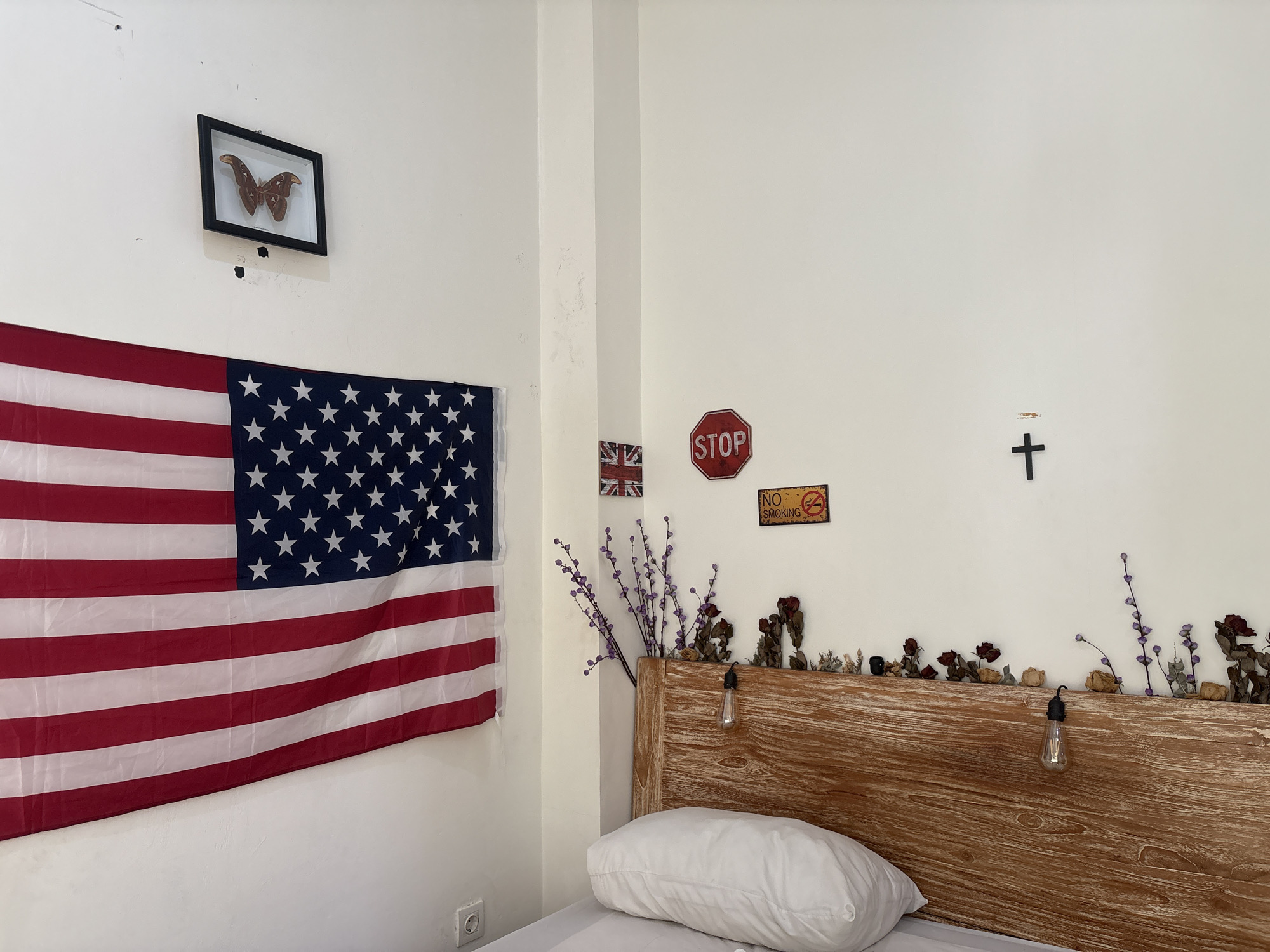 The image depicts a bedroom interior with a white wall as the primary background. On the left wall, an American flag is hung, and above it, a framed butterfly specimen is displayed. To the right of the flag, a small Union Jack flag is mounted. Further to the right, a red octagonal "STOP" sign is visible, followed by a yellow "NO SMOKING" sign. On the upper right side of the wall, a black Christian cross is mounted.
The foreground features a bed with a white pillow and a wooden headboard. The headboard is decorated with dried flowers and branches, and a single Edison-style light bulb is suspended above it. The room appears to be sparsely furnished, with a white wall-mounted electrical outlet visible on the lower left. The lighting suggests it is daytime, with natural light contributing to the overall brightness of the scene. The style suggests a bohemian or eclectic decor.