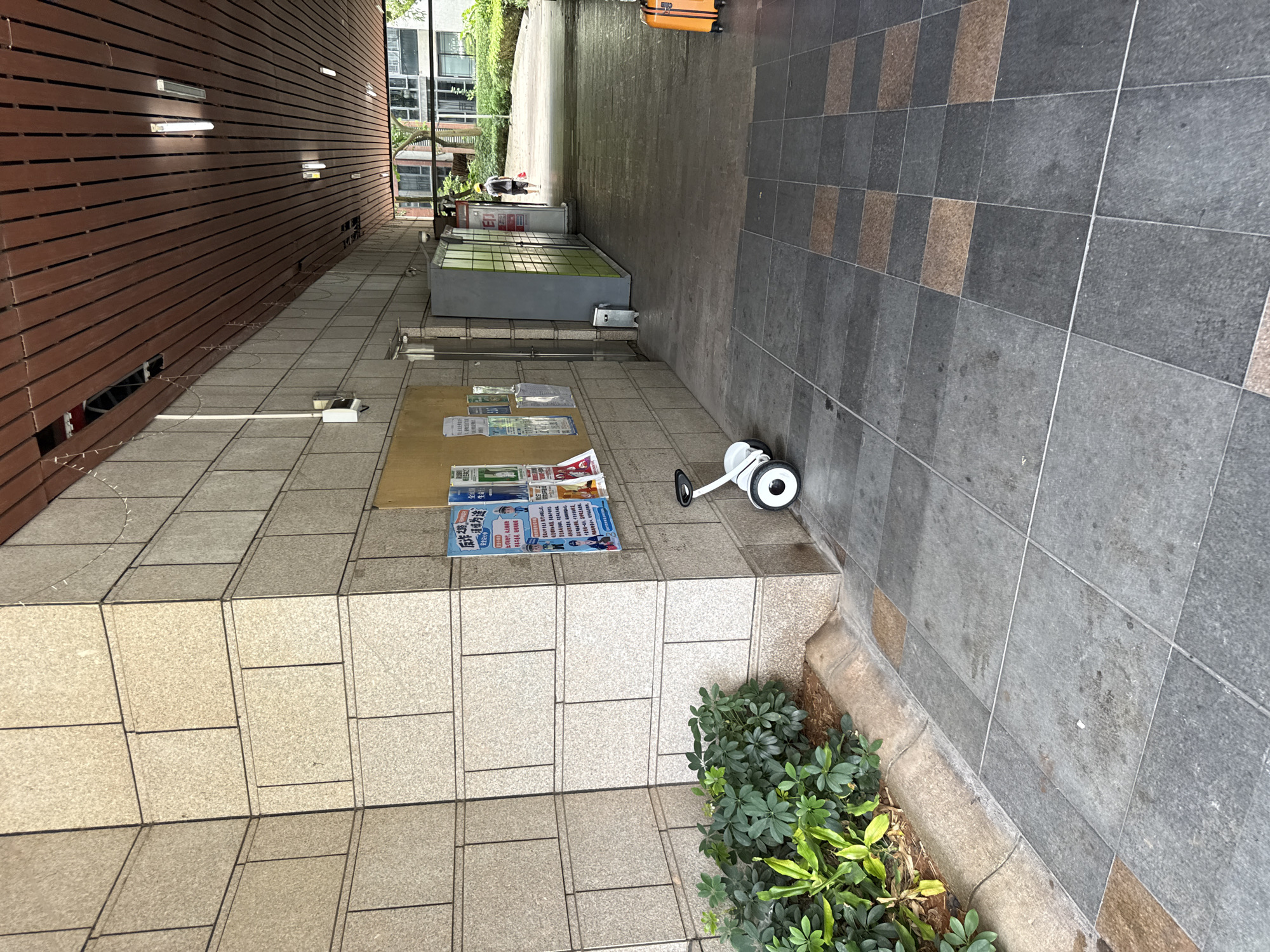 The image depicts an outdoor walkway in a modern urban environment, likely located in Haizhu District, China, characterized by its tiled pavement and a covered walkway with a wooden-slatted ceiling. The scene is captured from a ground-level perspective.
On the left, a wall constructed of large, light-colored granite-like tiles dominates the foreground. Adjacent to this wall is a dense patch of green foliage, including leafy plants and some drier ground cover. Attached to the tiled wall are several posters and notices, pinned to a corkboard.
A white self-balancing scooter (Segway) is positioned on the tiled walkway in the mid-ground, to the right of the plants. The walkway itself is paved with large, square tiles, with a pattern of darker grey and some reddish-brown tiles.
Further into the background, the walkway extends into a more open area with greenery and buildings visible. A person wearing a hard hat and carrying a red bag is walking away from the camera in the distance. To the right of the walkway, a row of green and grey lockers is visible, along with some trash receptacles. An orange suitcase is placed on the far right of the frame, near the edge of the walkway. The overall environment suggests a public area or thoroughfare within a complex, possibly residential or commercial.