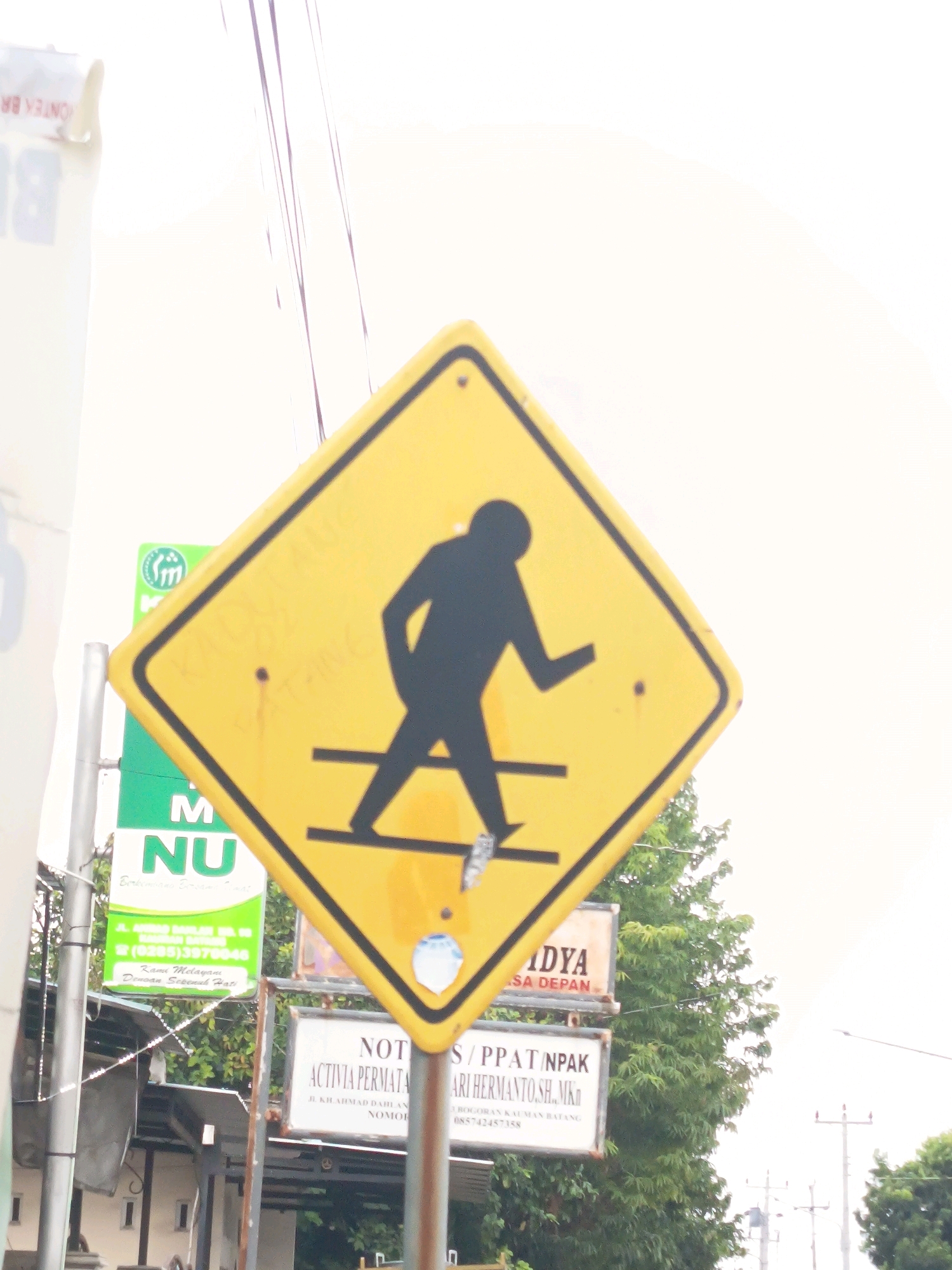 A pedestrian crossing sign is the primary focus of the image. The yellow, diamond-shaped sign depicts a black silhouette of a person walking across two parallel lines, indicating a pedestrian crossing. The sign is mounted on a pole, and there are other signs visible in the background. One green sign features the letters "M NU" along with text in Indonesian. Below this, another sign reads "NOTIS / PPAT / NPAK" followed by more Indonesian text. A large green tree is positioned to the right of the signs. The sky is overcast and bright. The scene appears to be on a street in an urban or suburban area.