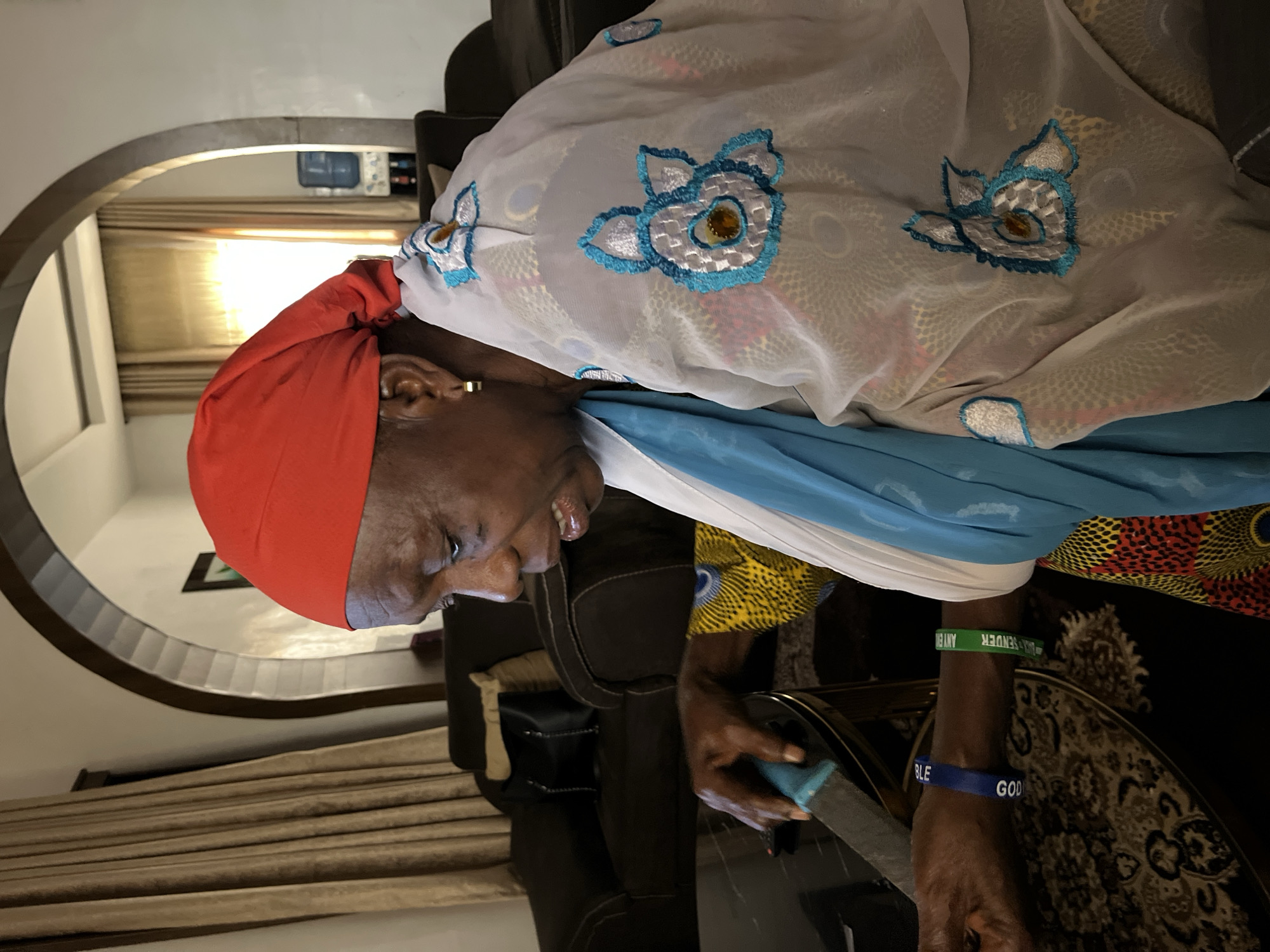 An image captures an individual indoors, rotated 90 degrees clockwise. The person, with dark skin, is wearing a red headscarf and a light, sheer garment adorned with blue and white embroidered floral patterns, featuring small gold-colored accents in the centers. Underneath, a bright blue fabric is visible, followed by a colorful patterned fabric with yellow, red, and black motifs. A gold-colored earring is visible in their right ear. Their mouth is slightly open, suggesting a relaxed expression or light speech, and they appear to be looking down and to their right.
On their right wrist, two silicone wristbands are worn: one green and one blue. The green band shows partial text, including "MAL SENDER" and "ANTEN". The blue band displays "BLE" and "GOD". The person's right hand is holding a light blue object against a dark surface.
The setting is an indoor space, likely a home, with an arched architectural feature visible in the background on the left. Curtains, possibly covering a window, are seen through the archway, indicating daytime lighting. A dark armchair or sofa back is behind the person, and a table with an intricate patterned surface is to their right. The overall atmosphere appears calm and candid. The attire and features are consistent with the provided location context of Jos, Nigeria.