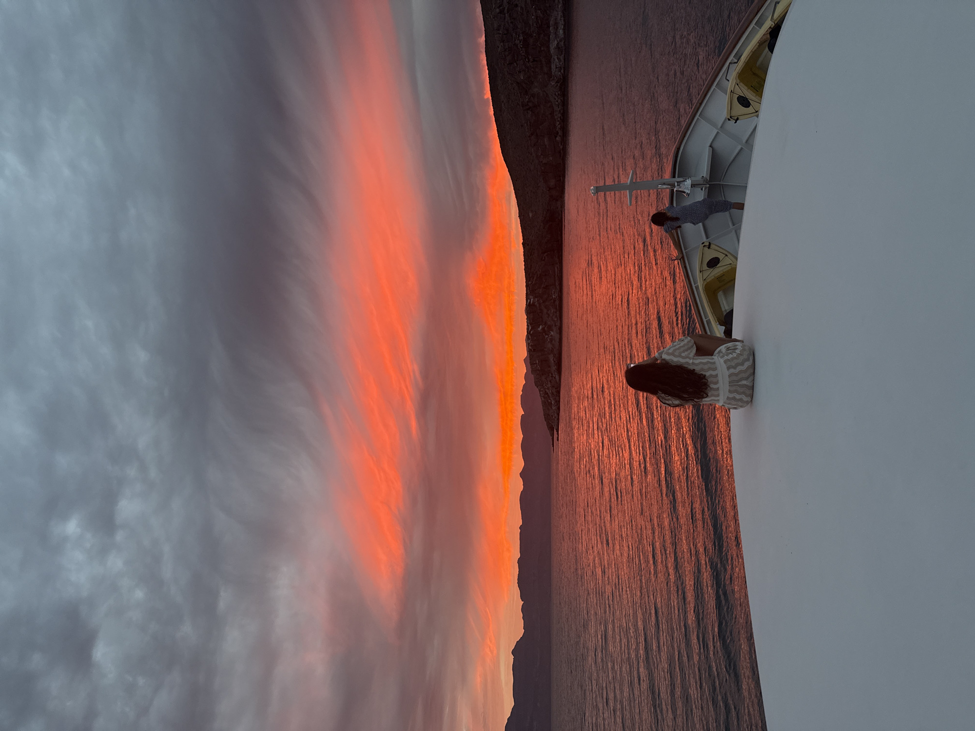The image depicts a sunset scene from the deck of a boat, possibly a cruise ship, in an unidentified location in Mexico. The sky is filled with grey clouds transitioning into vibrant orange and red hues near the horizon, which illuminates the water below. A dark silhouette of a mountainous coastline is visible along the horizon. Two people are on the deck. One is a woman with long brown hair, leaning against the white railing. The second person is a man standing further back, also near the railing. There are some safety boats on the side.