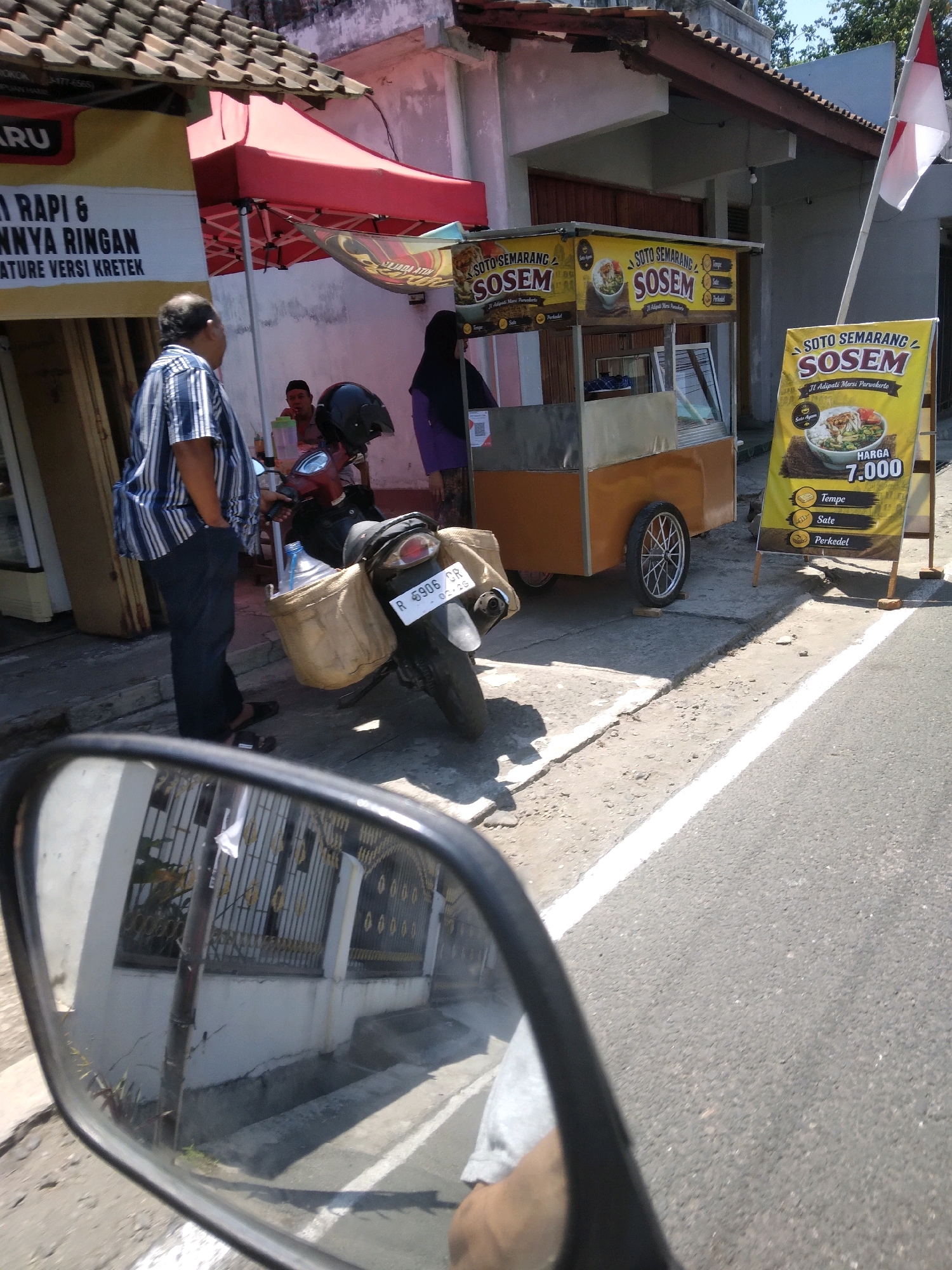 The image captures a street scene in Sokaraja, Indonesia, focused on a food stall selling "Soto Semarang". A mobile food cart, painted orange and silver, is prominently displayed, featuring signs advertising "SOTO SEMARANG SOSEM" and prices for items like "Tempe", "Sate", and "Perkedel". A large yellow banner with similar advertising and a price of "HARGA 7.000" stands beside the cart. A person in a blue and white striped shirt and dark pants stands near the cart, looking away from the camera. A motorcycle with a large woven basket on the back is parked behind the person. In the background, a red awning covers part of the stall area. The scene takes place on a paved street with a white line marking the edge. The reflection in a motorcycle's rearview mirror shows a building with barred windows and a patterned fence. Another person wearing a dark headscarf is partially visible behind the food cart. The overall environment suggests a busy roadside food vendor operation.