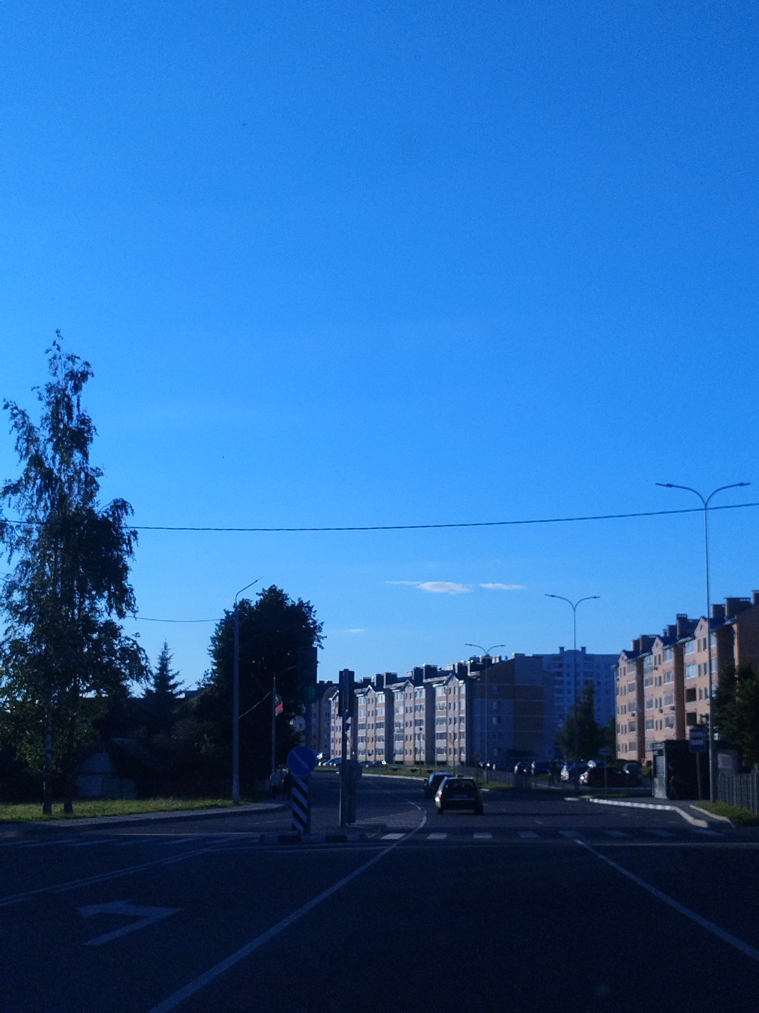 A daytime, eye-level, outdoor, wide shot captures a clear blue sky over an urban landscape with a road in the foreground and multi-story buildings in the background. A thin, dark horizontal line, likely a power line, stretches across the sky. On the left side of the image, a tall, slender tree with dark green foliage stands prominently. To the right, a series of uniform, light-colored apartment buildings with dark roofs and numerous windows form a residential block. A black car is visible driving on the road, which is marked with white lane lines and a directional arrow. Traffic signals and streetlights are also present. The road surface appears dark and wet, suggesting recent rain or a reflection from the sky. A pedestrian crossing is visible on the road. The scene conveys a sense of urban daily life on a bright day.