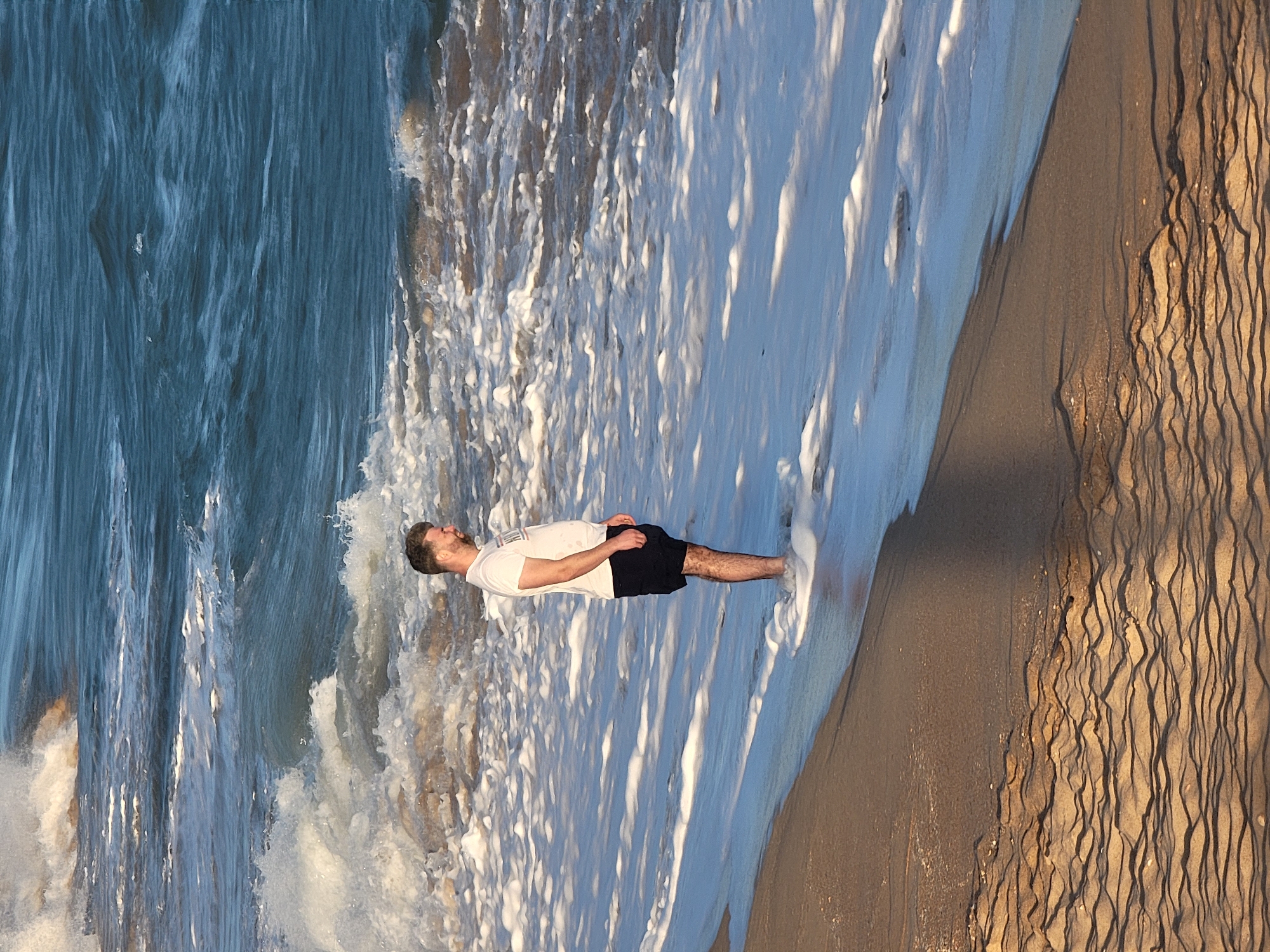 A man is standing in the shallow ocean water on a beach in Quarteira, Portugal. He is wearing a white t-shirt and dark shorts. The water is breaking around his legs, and a wave is washing over the sandy shore. The sun appears to be low in the sky, casting long shadows and illuminating the sand with a golden light. The sky is clear and the weather appears to be warm and pleasant. The scene evokes a sense of peace and tranquility.