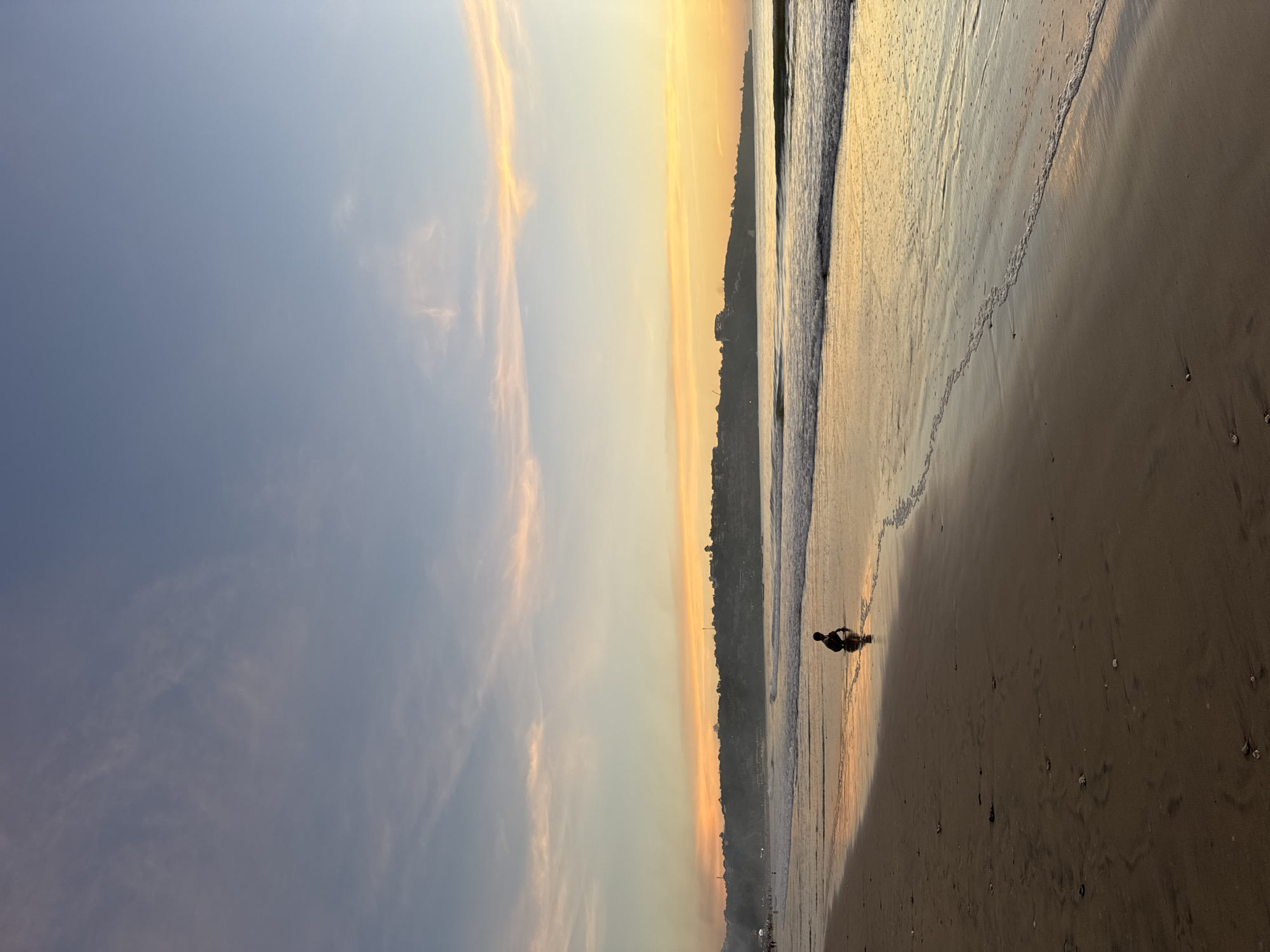 A beach scene at sunset in Kuta Selatan, Indonesia. The sky transitions from blue to orange near the horizon. A coastline with buildings is visible. Waves break on the shore. A person stands near the water's edge. The sand is wet.
