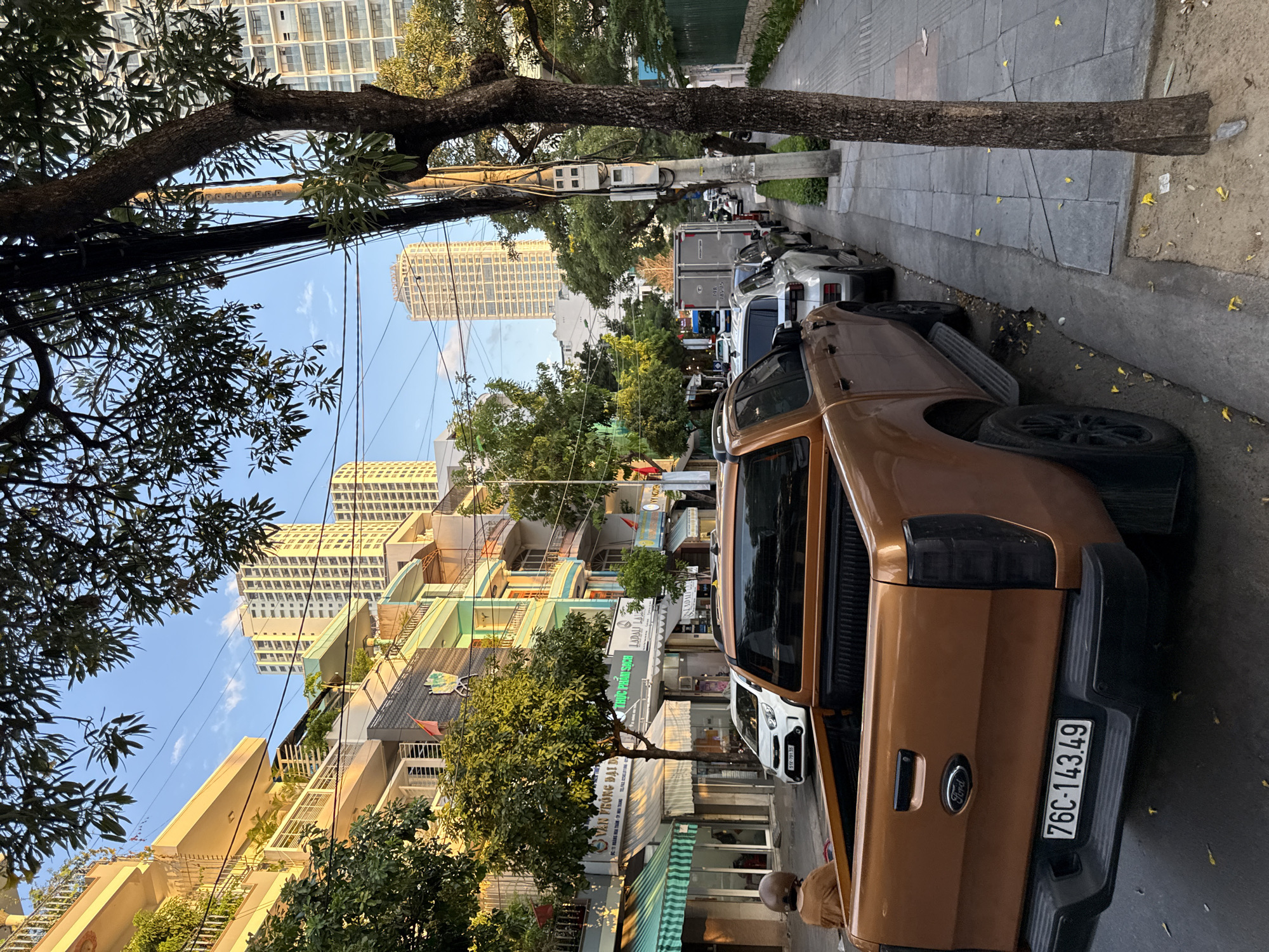 The image depicts a street scene in Nha Trang, Vietnam, likely during late afternoon or early evening, given the golden hue of the sunlight. The perspective is from behind a brown Ford pickup truck parked on the right side of the street, facing away from the camera. The truck has a license plate that reads "76C-143.49".
To the left of the truck, a sidewalk runs parallel to the street, bordered by a row of multi-story buildings. These buildings exhibit a mix of architectural styles, with some featuring balconies, awnings, and decorative facades. Several trees line the sidewalk, their leaves casting dappled shadows.
Further down the street, several other vehicles are parked, including a white hatchback and a white SUV. Tall, modern apartment buildings are visible in the background, indicating a developing urban environment. A person wearing a helmet and a brown jacket is seated on the edge of the sidewalk near the rear of the pickup truck, facing away from the camera.
The street appears to be a moderately busy thoroughfare, with a sidewalk and streetlights present. Power lines and utility poles are visible, contributing to the urban landscape. The overall impression is that of a typical city street in Vietnam, with a blend of older and newer construction, residential and commercial properties, and the presence of vehicles and pedestrians.