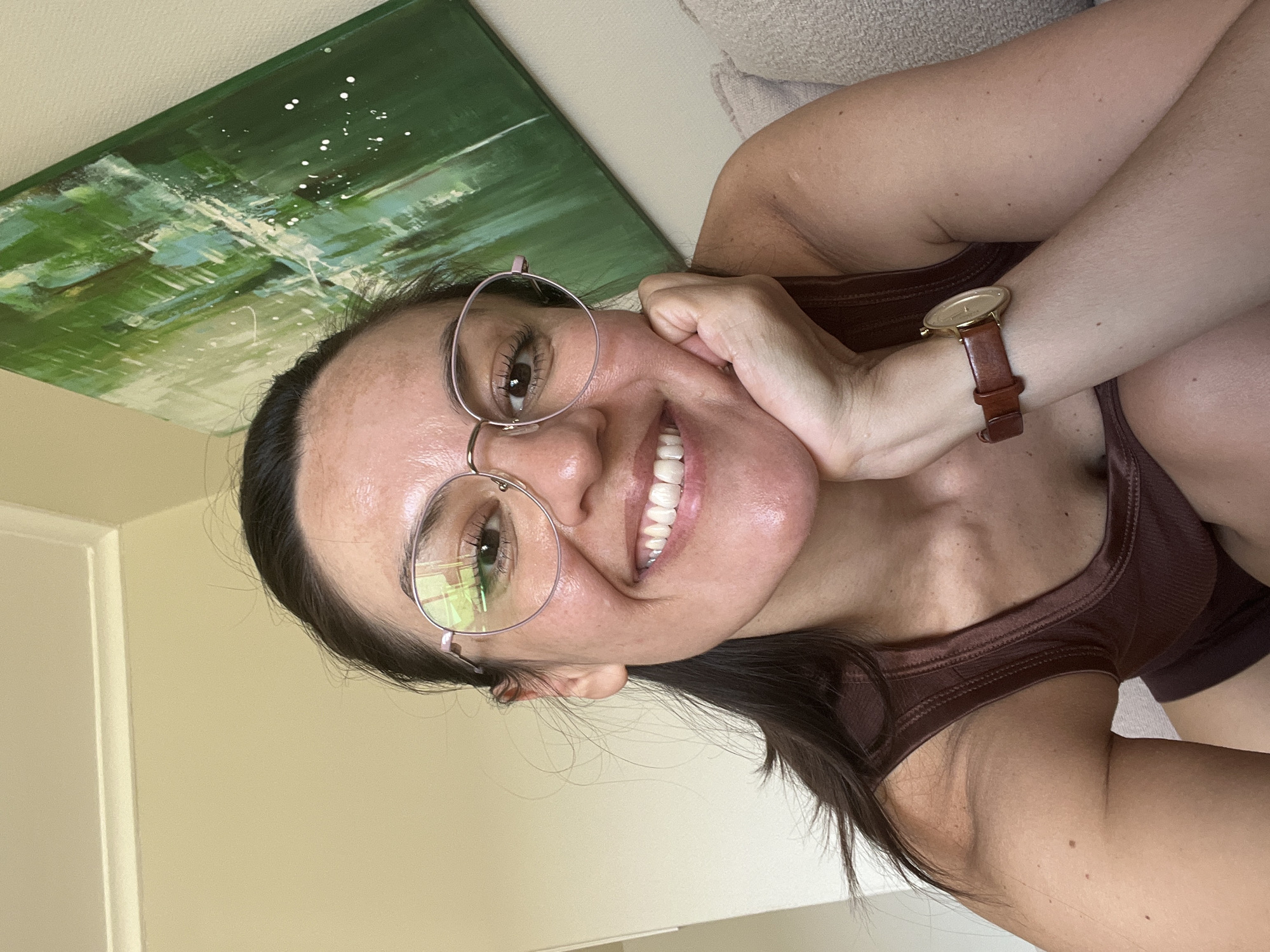 A woman with dark hair, wearing glasses and a brown tank top, is smiling and posing for a selfie. She is wearing a watch with a brown leather strap. Behind her is an abstract green and white painting. The wall appears to be light yellow. The location is in Herlev, Denmark.