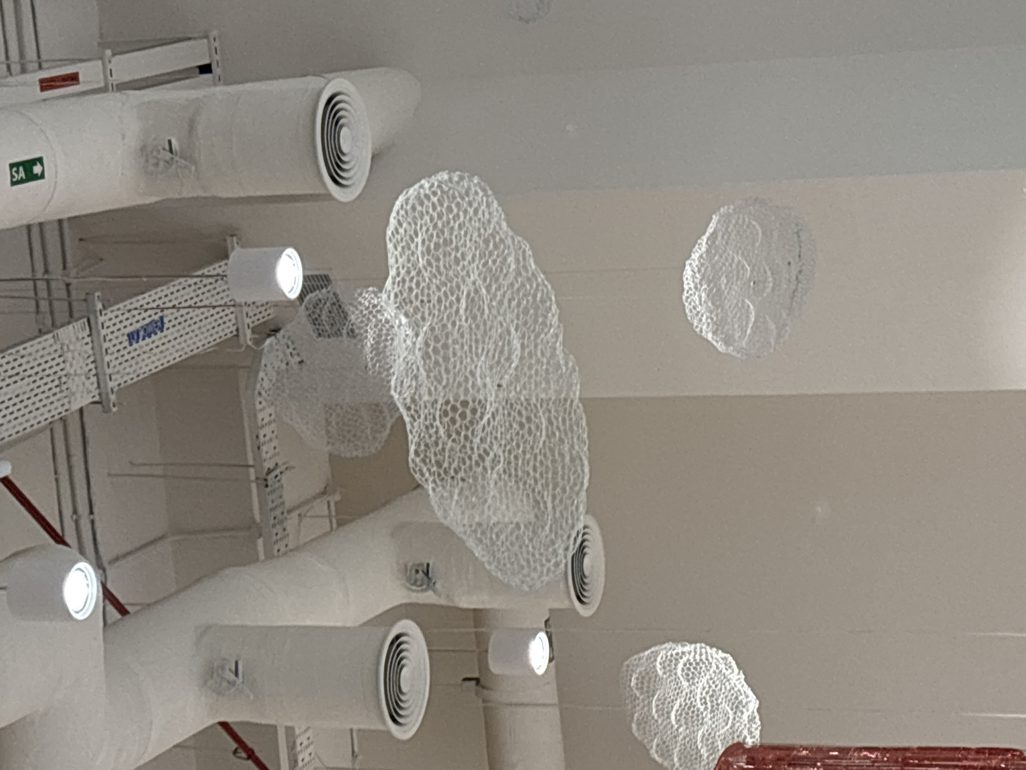 The image shows an interior view of a building's ceiling area, featuring a complex network of white ductwork, exposed conduits, and circular air vents. Hanging from the ceiling are several large, white, mesh-like structures resembling clouds. One prominent cloud structure is centrally located, with smaller ones suspended at various heights and positions. White cylindrical lights are also visible, attached to the ceiling. A red pipe runs diagonally across the upper portion of the image. The overall aesthetic suggests a modern commercial or public space. The specific location within Dubai, United Arab Emirates cannot be determined from the visual information provided. There are no people or discernible actions depicted in the scene.