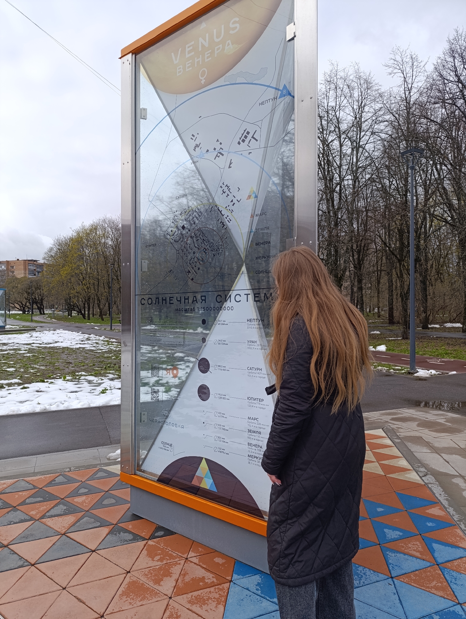 A person with long, light brown hair, wearing a black quilted jacket and dark pants, stands with their back to the camera, intently viewing a large, vertical outdoor display panel. The scene is an overcast daytime in what appears to be late winter or early spring, given the bare trees and patches of melting snow on the ground.
The display panel is titled "VENUS ВЕНЕРА" at the top and prominently features "СОЛНЕЧНАЯ СИСТЕМА" (Solar System) with a scale of "1:1500000000". It presents information about the planets, including their orbital distances in kilometers and their scaled distances "в городе" (in the city), indicating a scaled model of the Solar System laid out within the location. Visible planet names include "МЕРКУРИЙ" (Mercury), "ВЕНЕРА" (Venus), "ЗЕМЛЯ" (Earth), "МАРС" (Mars), "ЮПИТЕР" (Jupiter), "САТУРН" (Saturn), "УРАН" (Uranus), and "НЕПТУН" (Neptune), along with "СОЛНЦЕ" (Sun). For example, Earth's orbit is listed as "150 млн км" (150 million km) and "100 м в городе" (100 m in the city). The text "ЧЕРНОГОЛОВКА" is visible near the bottom of the panel, identifying the location as Chernogolovka, Russia.
The ground around the display is paved with an eye-catching pattern of interlocking terracotta, gray, and blue triangular tiles. In the background, there is a paved path or road, a red bicycle lane, and several leafless trees. A building can be seen in the far distance. QR codes are also visible on the display, likely providing additional information.