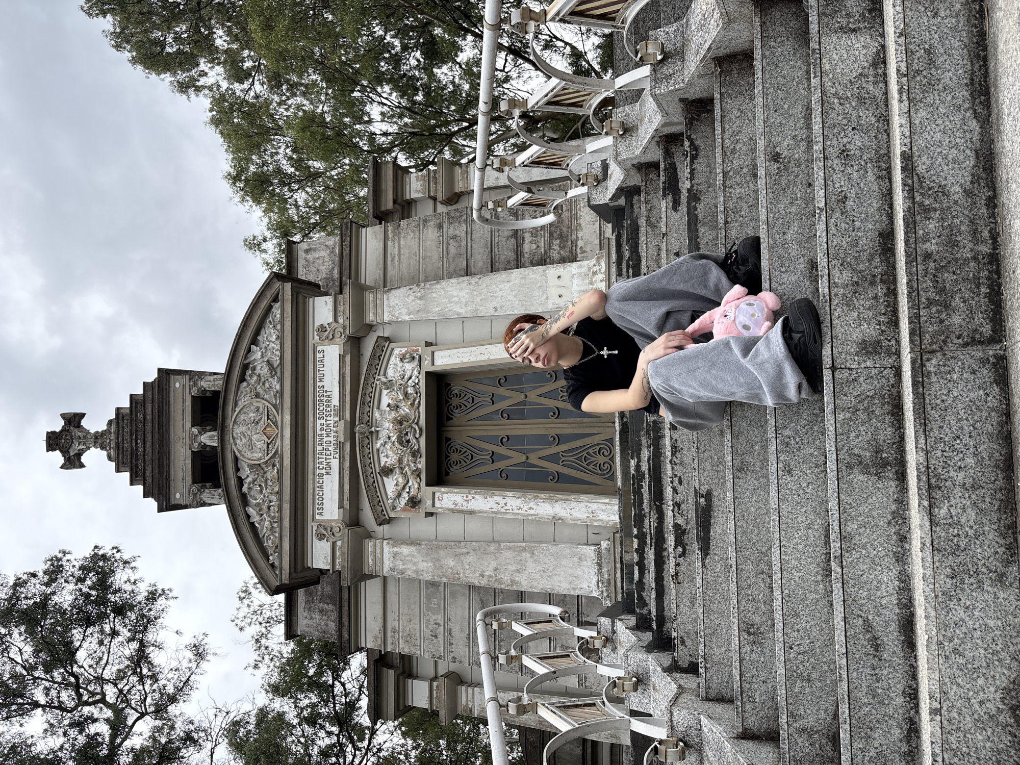 An individual sits on the weathered granite steps of the "ASSOCIACIO CATALANA DE SOCORS MUTUAS MONTEPIO MONTSERRAT" mausoleum located in Recoleta Cemetery, Buenos Aires, Argentina. The mausoleum, constructed of light-colored stone, features an ornate facade with decorative carvings, pillars, a central double door, and a large stone cross topping its multi-tiered roof. The sky overhead is overcast, and sparse trees with green foliage are visible on either side of the structure.
The subject has reddish-brown hair, wears a dark short-sleeved top, grey sweatpants, and black sneakers. A silver-colored cross pendant hangs from their neck, and tattoos are visible on their left hand. They are seated on the third visible step from the top, with their left hand covering their eyes, and their head tilted slightly upward and to the left. Their right hand holds a small, pink, plush character resembling My Melody, which rests on their lap. The granite steps and mausoleum show visible signs of weathering and dark staining.