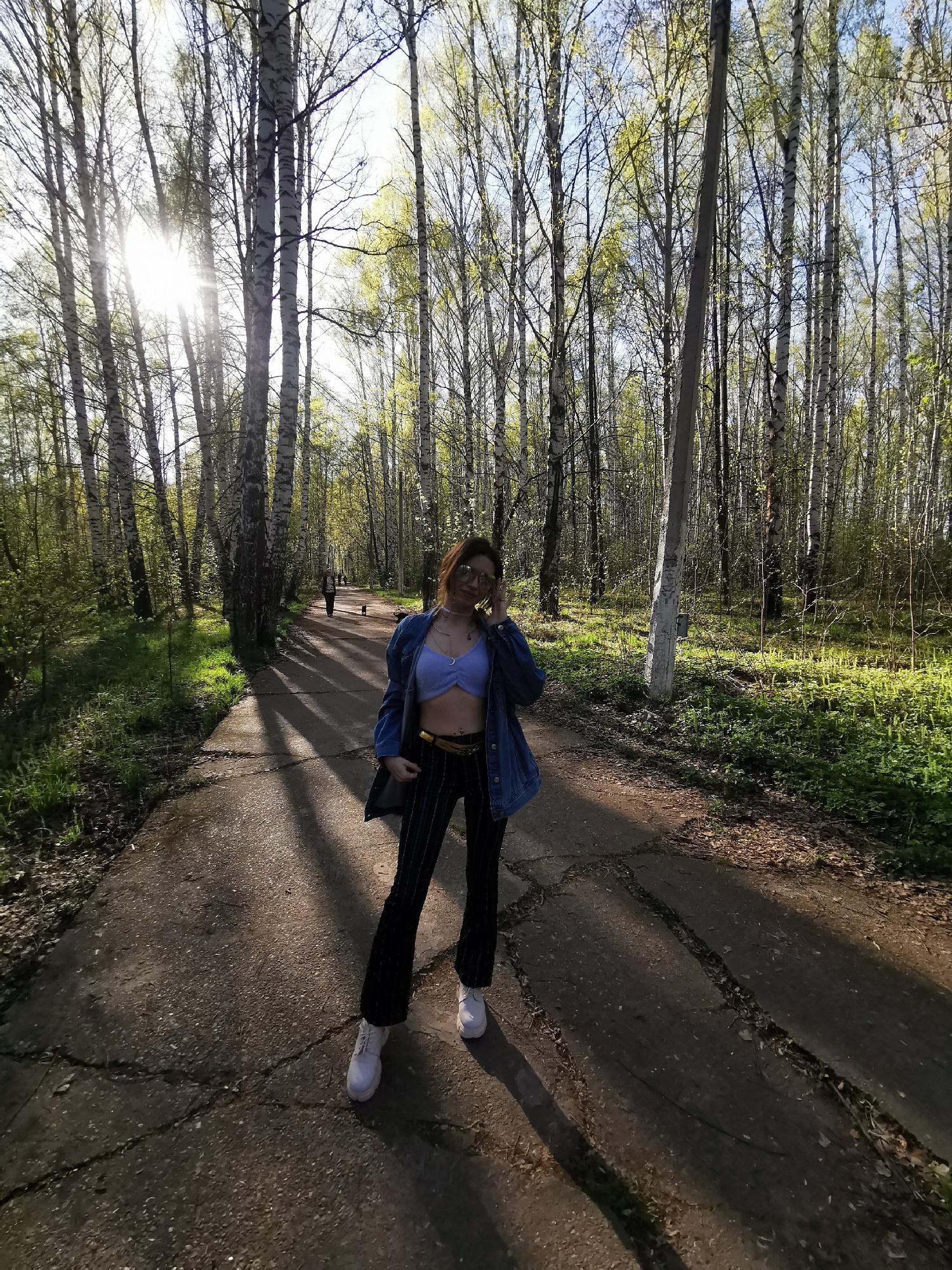 The image captures a person standing on a cracked concrete path within a birch tree forest. The individual is wearing a blue bralette, a denim jacket, pinstripe pants, and white sneakers. Sunlight streams through the trees, casting shadows on the path. In the distance, another person is walking with a dog. The location appears to be a park or forested area within the city.