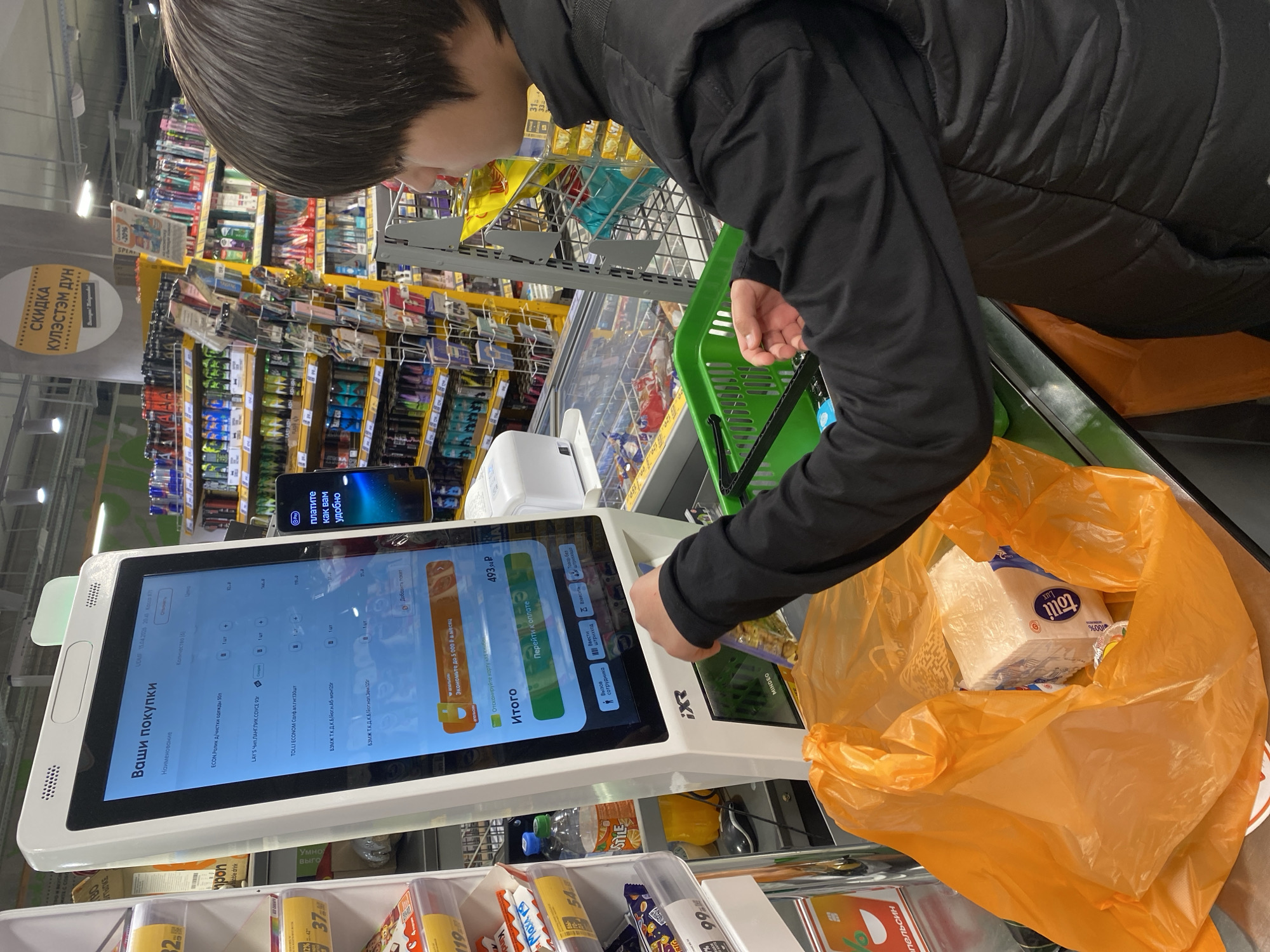 Here's a forensic description of the image:
**Scene:** Interior of a store, specifically at a self-checkout station.
**Subject:** A person with short, dark hair, wearing a black long-sleeved shirt and a black vest. They are operating the self-checkout machine.
**Objects:**
* Self-checkout machine: Displays the items being purchased ("Ваши покупки"). The total is 493.93P. There are options to pay, call for assistance.
* Shopping cart: Contains a green basket and an orange plastic bag. The bag holds a package of "Tolli Lux" tissues.
* Merchandise: Shelves in the background are stocked with various items. Candies, gums and confectionaries are visible at the checkout lane.
* Credit card reader.
* Smartphone glued to the top of the self-checkout machine with the message: "Платите как вам удобно" ("Pay as you wish").
**Actions:**
* The person is using the self-checkout machine to scan their items.
**Environment:**
* Well-lit store with aisles of merchandise in the background.
* A sign is visible in the background reading, "СКИДКА КУЛЭСТЭМ ДУН" ("Discount Kulestem Dun").
Based on the OCR text and sign, the location is inferred to be a store in Russia, potentially in the city of Votkinsk.