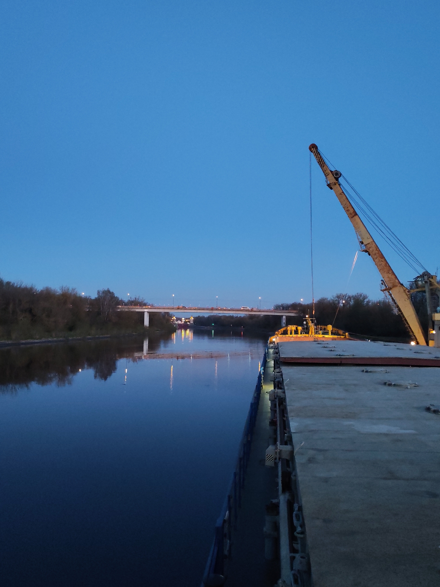 A medium shot captures a broad barge equipped with a sizable crane situated on the Yakhroma River in Russia. The crane, positioned on the starboard side of the barge, is partially visible. The barge deck is clear. Across the river is a bridge with visible car traffic moving over it. Both banks of the river are lined with trees. The scene is captured during dusk, with lights beginning to illuminate the surroundings, particularly on the bridge and on the barge itself. The sky above is a clear blue.