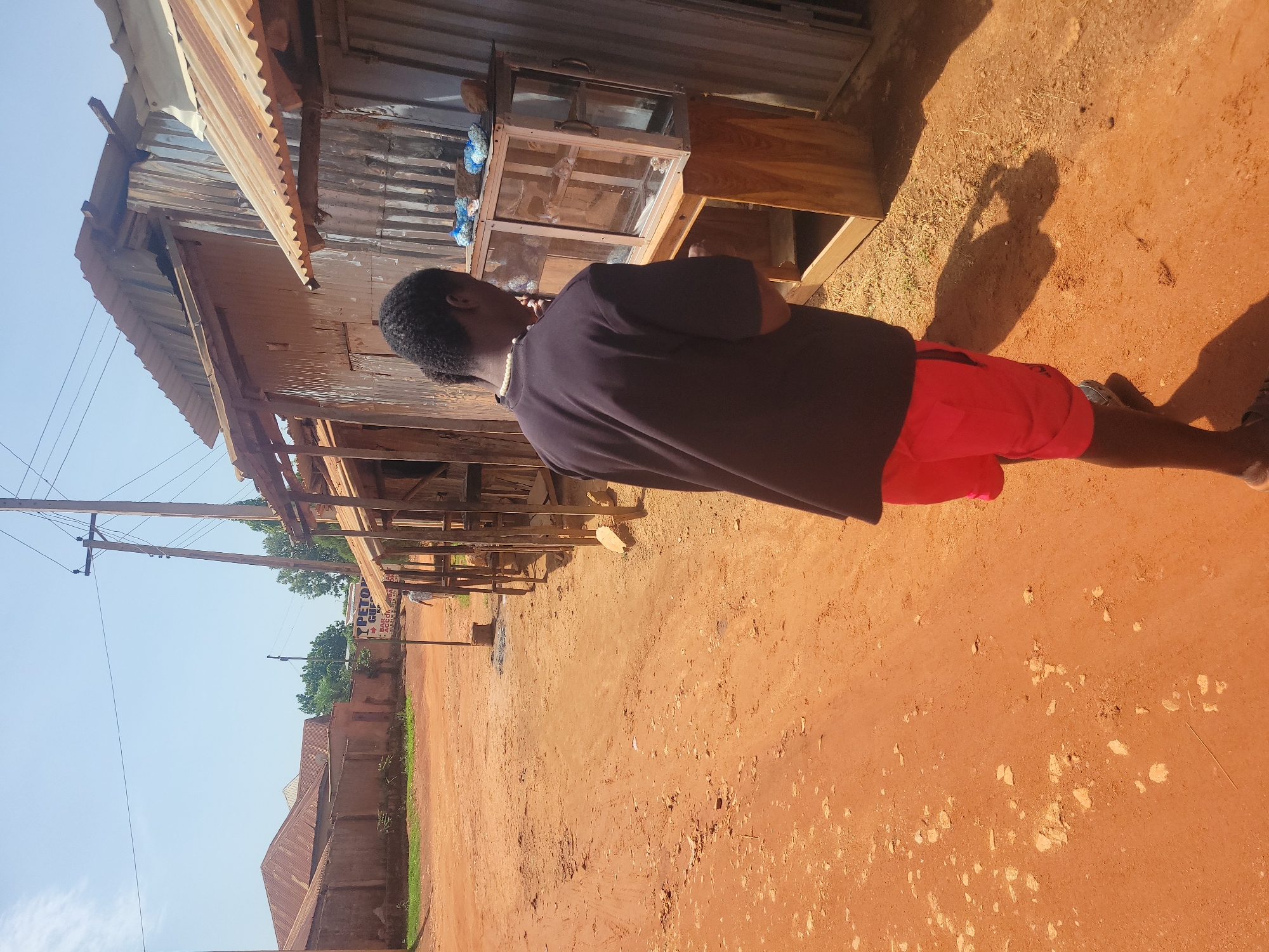 A full shot captures a person in a red shirt and black shorts, with black hair, standing on a dirt road under a clear sky. The person is looking at a glass display case on a wooden stand, which is next to a building constructed with corrugated metal and wood. The scene is set in Jos, Nigeria, with several buildings featuring tin roofs visible in the background, along with a utility pole and overhead wires. The ground is covered in reddish-brown dirt with scattered rocks and sparse vegetation.