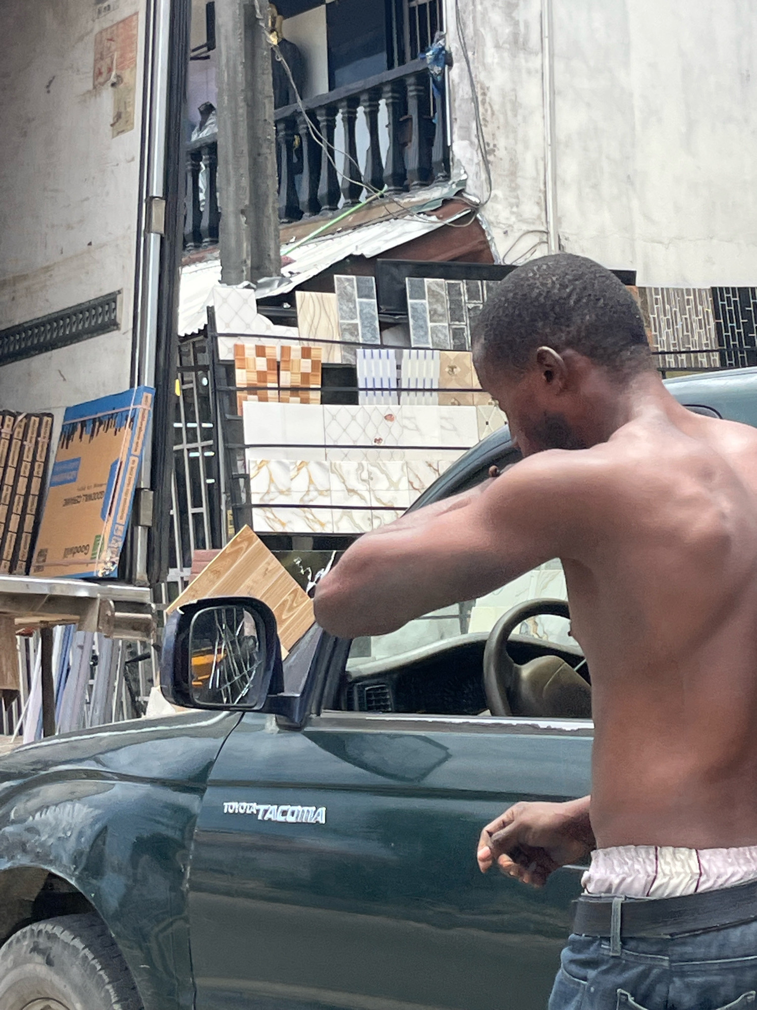 An individual, shirtless and wearing jeans and white striped boxer briefs, is seen from the side in Ado, Nigeria. The person is leaning towards a dark green Toyota Tacoma truck, with their right arm extended towards the vehicle's side mirror. A light brown rectangular object, possibly a tile, is being held in their right hand. The background features a building with an upper-level balcony and a display of various tiles. The scene appears to be on a street with building materials.