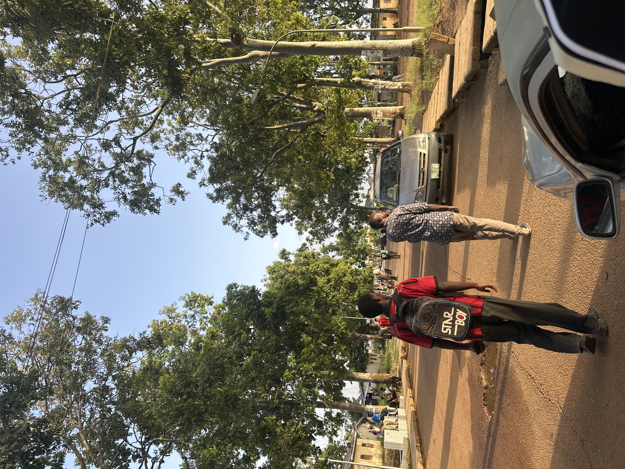This image depicts an outdoor scene in Rafin Par, Nigeria. The time of day appears to be late afternoon, with long shadows cast on the asphalt road, suggesting the sun is low in the sky. The weather seems to be clear and sunny.
In the foreground, two men are walking on the road. The man on the left is wearing a patterned short-sleeved shirt and light-colored pants. The man on the right is wearing a red short-sleeved shirt and dark pants, and he has a black backpack with the text "STAR BOY" printed on it. There is also a silver car parked on the right side of the frame, with its side mirror visible.
The background features a tree-lined street with large trees casting shade over the road. Beyond the trees, buildings are visible, including some with visible rooftops and windows. Other people can be seen in the background, suggesting a public or commercial area. The road itself is paved with asphalt and has a curb along the side. The overall setting appears to be a typical street scene in a Nigerian town or city.