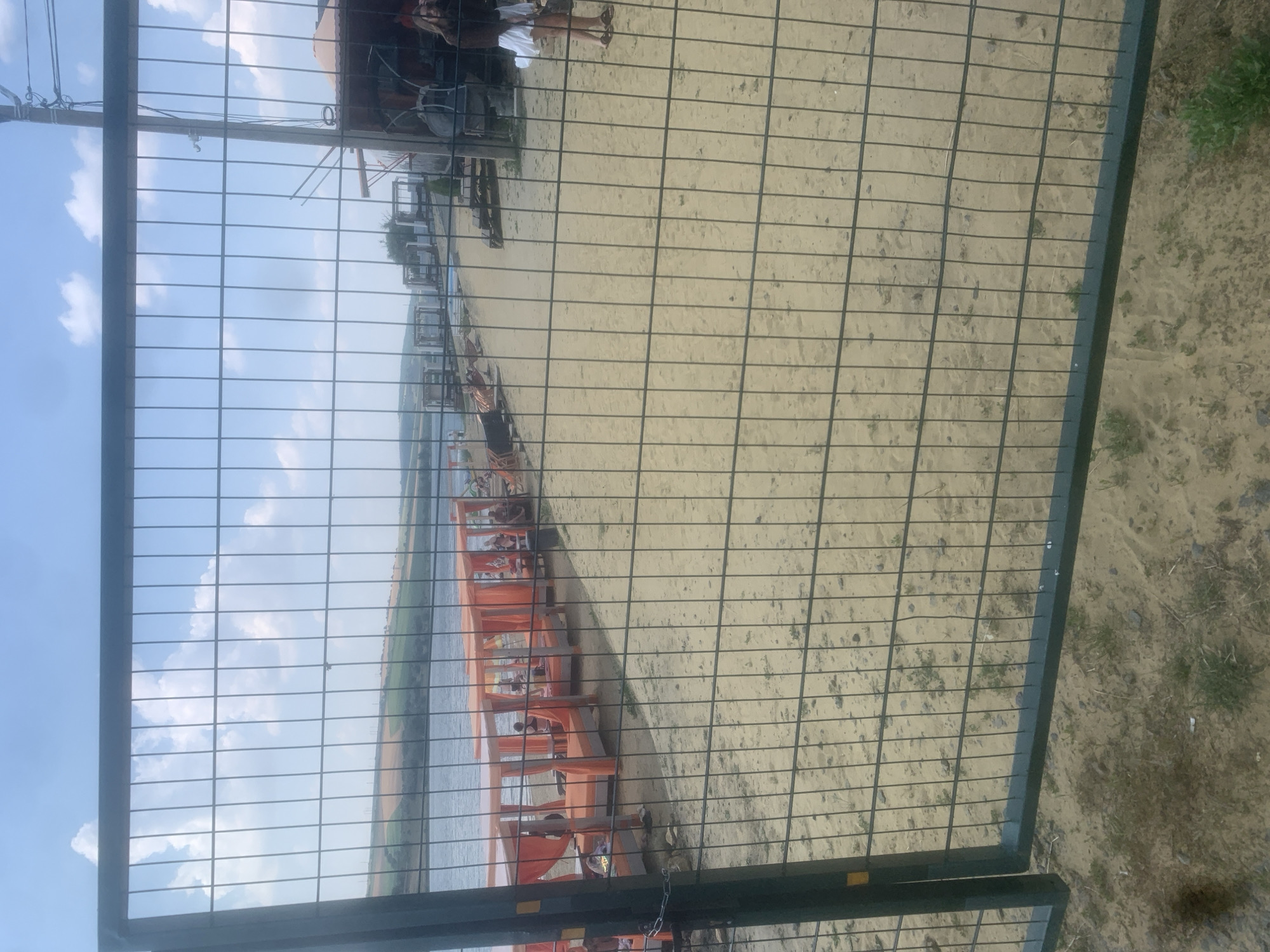 The image shows a sandy area with a body of water to the left, viewed through a wire fence. Several orange structures, possibly beach cabanas or tents, are set up on the sand near the water. People are visible within and around these structures, engaged in various activities. The sky is blue with some clouds, suggesting fair weather. The scene appears to be a beach or lakeside recreational area, likely during the daytime given the lighting. The sand is light brown, and there are some sparse patches of greenery. The overall impression is one of a relaxed outdoor setting.