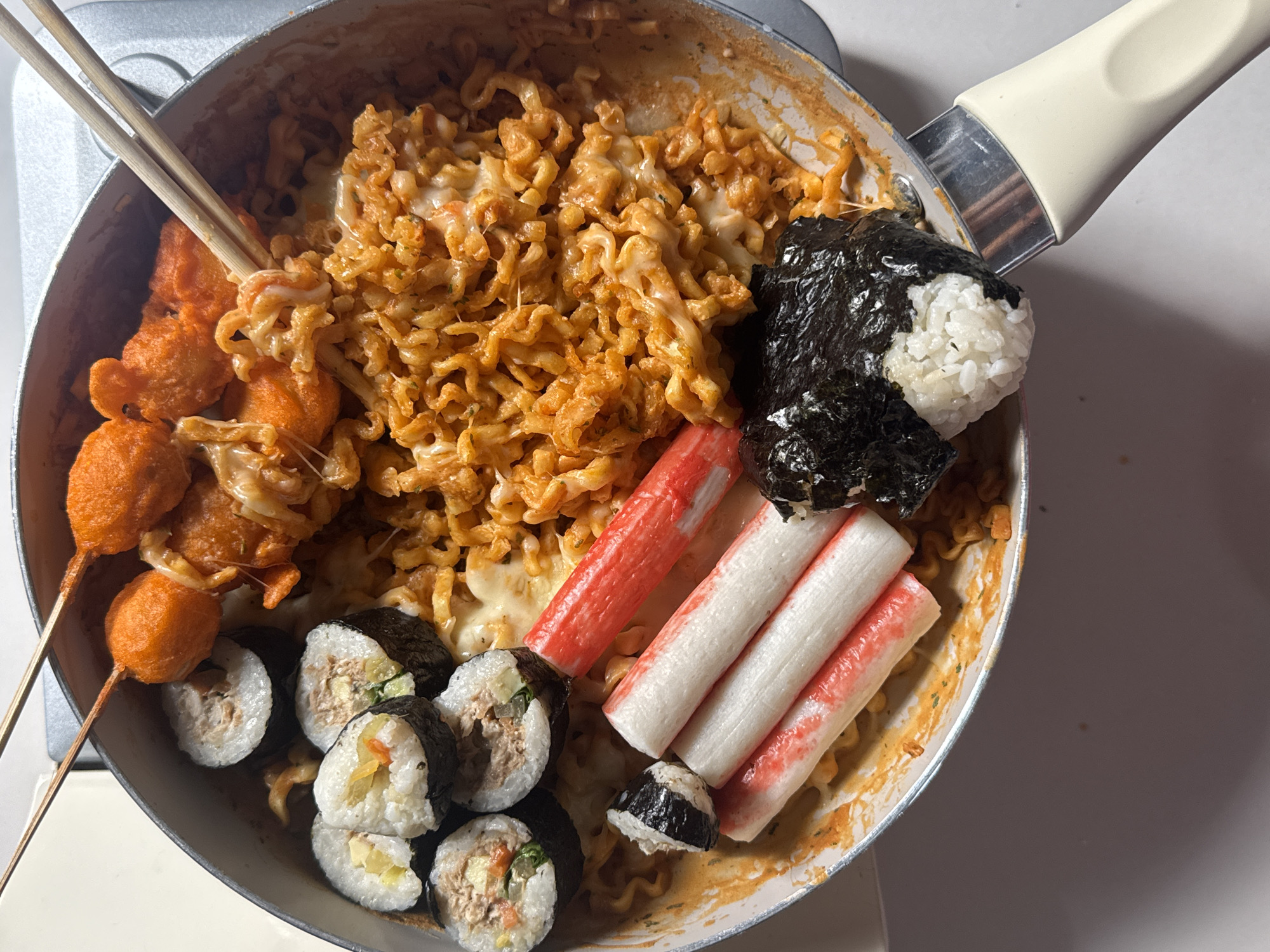 The image displays a close-up, top-down view of a cooking pot filled with a meal. The pot contains a generous portion of spicy instant noodles, mixed with melted cheese, and several other food items arranged around the noodles. These items include what appear to be sushi rolls, some sort of fried orange balls on skewers, imitation crab sticks, and a small portion of rice wrapped in seaweed. Chopsticks are placed on top of the noodles, suggesting the food is ready to be eaten. The setting appears to be a kitchen, with a stovetop visible in the background and a plain white surface below the pot. There is no indication of any specific location within Davao City, Philippines; the image solely focuses on the food in the pot.