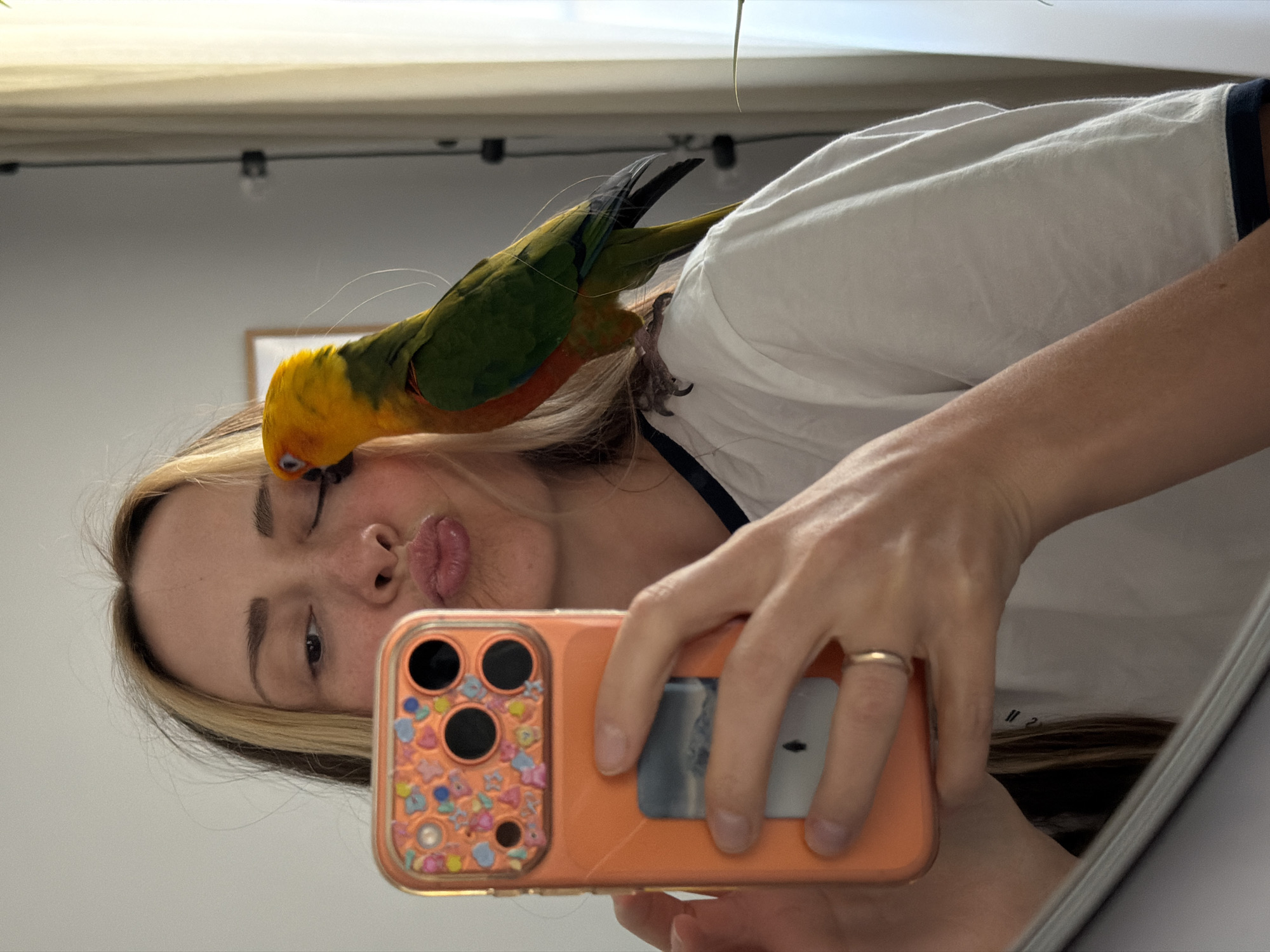 A young woman is taking a selfie in a mirror. A bird, identified as a sun conure, is perched on her head. The woman has blonde hair and is wearing a white t-shirt with navy trim. She is holding an orange cell phone, which features a clear square insert on the back. The location appears to be inside a room, indicated by the ceiling fixture in the background. There is no evidence to confirm that the location is in the city of Khimki, Russia.