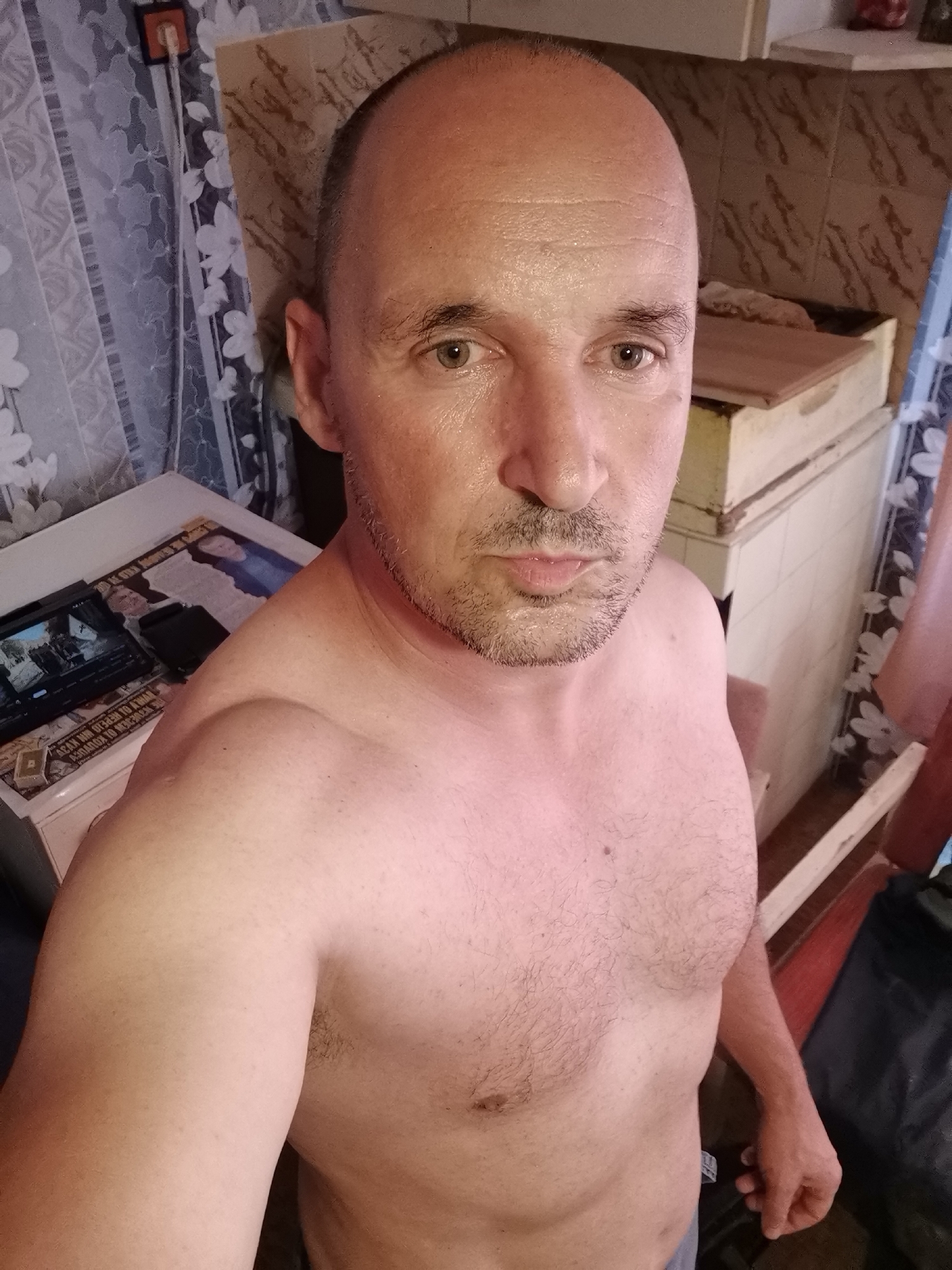 A shirtless man takes a selfie indoors. He is middle-aged, bald or has very short hair, with a light stubble beard and mustache. His skin appears slightly sweaty or glistening, and he has a neutral expression, looking directly at the camera. His chest shows some light body hair.
The background on the left features patterned curtains, possibly with white floral designs on a light blue or gray fabric. Below the curtains is a white table or cabinet. On this surface, a tablet or small screen displays a video, and next to it lies a black object. A newspaper or magazine is spread out, showing images and inverted text, including phrases in Cyrillic such as "О ЧЕМ НАПИСАТЬ" (WHAT TO WRITE ABOUT), "НА ЧТО НАДЕЯТЬСЯ" (WHAT TO HOPE FOR), and "ВЕЛИКИЙ ИСТОРИК К. МАРКС" (GREAT HISTORIAN K. MARX).
The background on the right reveals a different part of the room, possibly a storage unit or rustic counter with shelves, made of light-colored wood or painted material. Behind this unit, a wall with patterned tiles or wallpaper is visible. The lighting is artificial and warm, consistent with an indoor setting.