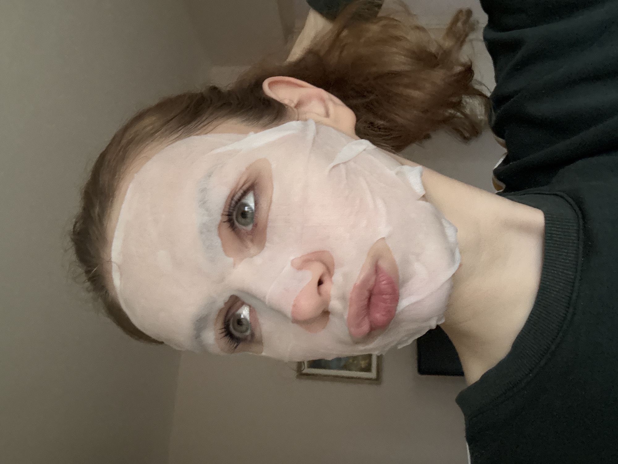 An individual, identified as a young adult female, is depicted indoors in Belgrade, Serbia. She is presented in a medium close-up, facing slightly left from the viewer's perspective, with her gaze directed upwards and to the left. Her face is predominantly covered by a white, translucent sheet mask, which features precisely cut openings for her eyes, nostrils, and mouth. Her eyes are light in color, and her eyelashes are distinctly visible. Her lips are pursed into a specific expression. Her hair is light brown to auburn, appears to be pulled back, and is visible on the right side of her head and extending behind her left ear. She is attired in a dark green long-sleeved garment with a ribbed collar. A portion of her left hand is visible on the right side of her head, appearing to interact with her hair. The background consists primarily of a plain, light-colored wall. A framed picture, featuring a dark frame, is mounted on the wall in the mid-left background. A dark, indistinct object is partially visible in the lower-mid background.
