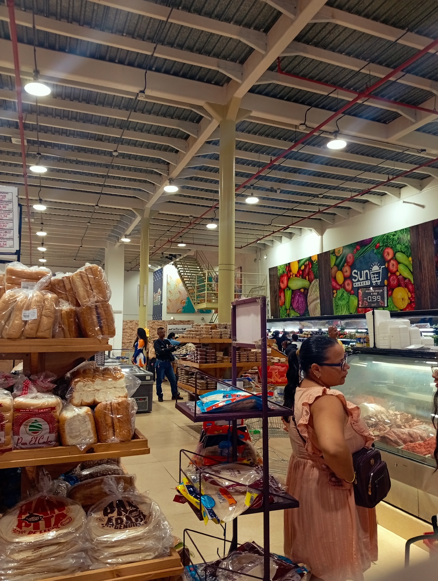 The image depicts an interior view of a supermarket in Juan Griego, Venezuela. The scene is characterized by rows of shelves stocked with various food items, primarily bread and tortillas, packaged in plastic. In the foreground, a woman wearing a light pink dress and glasses is observed from the side, looking towards a refrigerated display case filled with meat products. Behind her, other shoppers and employees are visible amidst the aisles. The supermarket features a high ceiling with exposed beams and industrial lighting. A large mural featuring colorful produce and the "Sun Market" logo is prominent on the back wall. Stairs are visible in the background, suggesting a multi-level store. The overall impression is that of a busy retail environment.