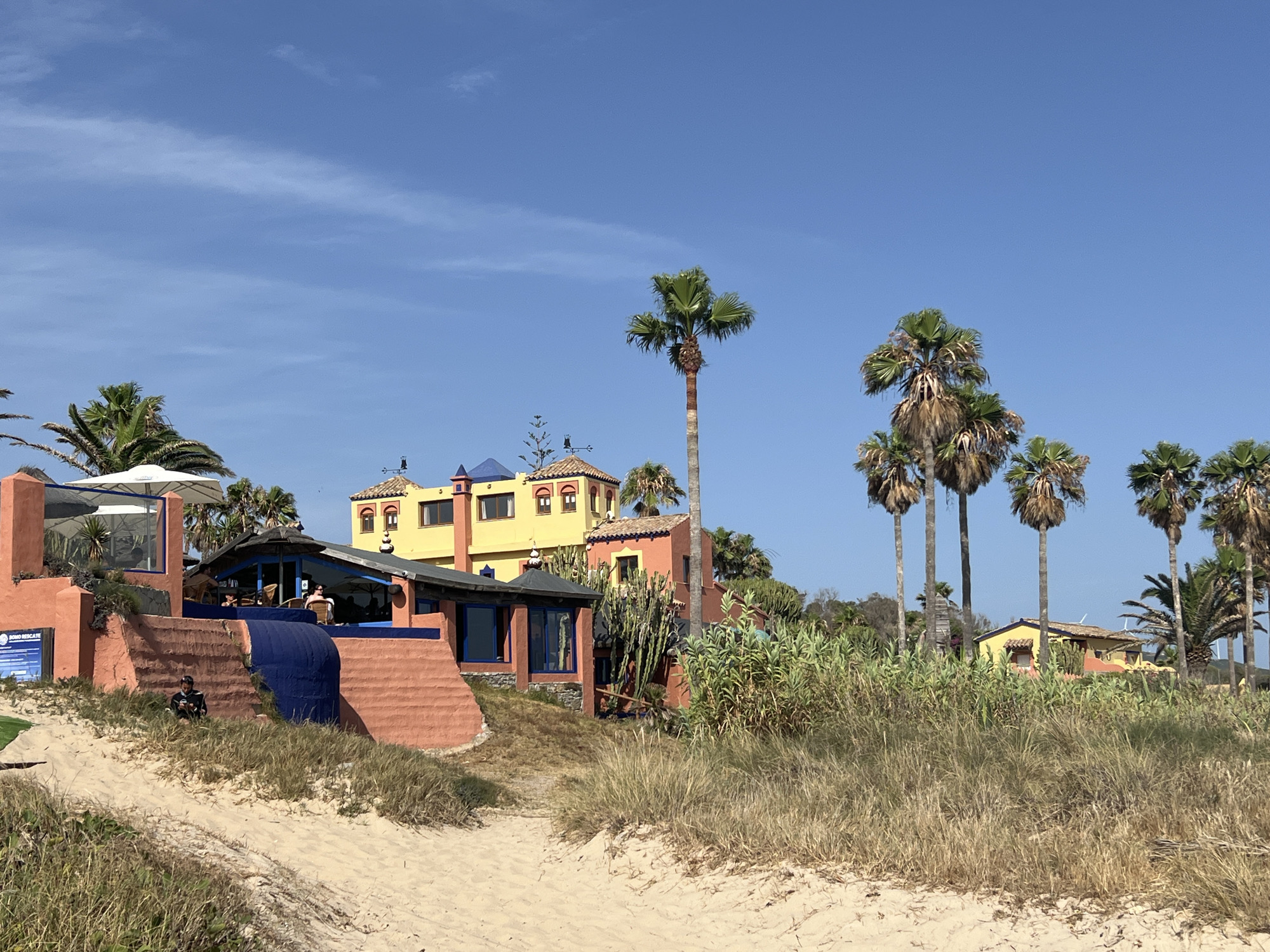 This image depicts a coastal scene in Tarifa, Spain, featuring a colorful building complex with red and yellow stucco walls, set against a clear blue sky. The architecture incorporates arched windows and tiled roofs, suggesting a Mediterranean influence. Palm trees are abundant throughout the scene, framing the buildings and dotting the landscape. In the foreground, sand dunes covered in dry grass lead towards the structures. A person is visible sitting on the sandy slope in front of one of the buildings, facing a smartphone. Another structure, possibly a restaurant or cafe, has outdoor seating with umbrellas and people seated at tables. The overall impression is one of a sunny, relaxed day at a beachside location.
