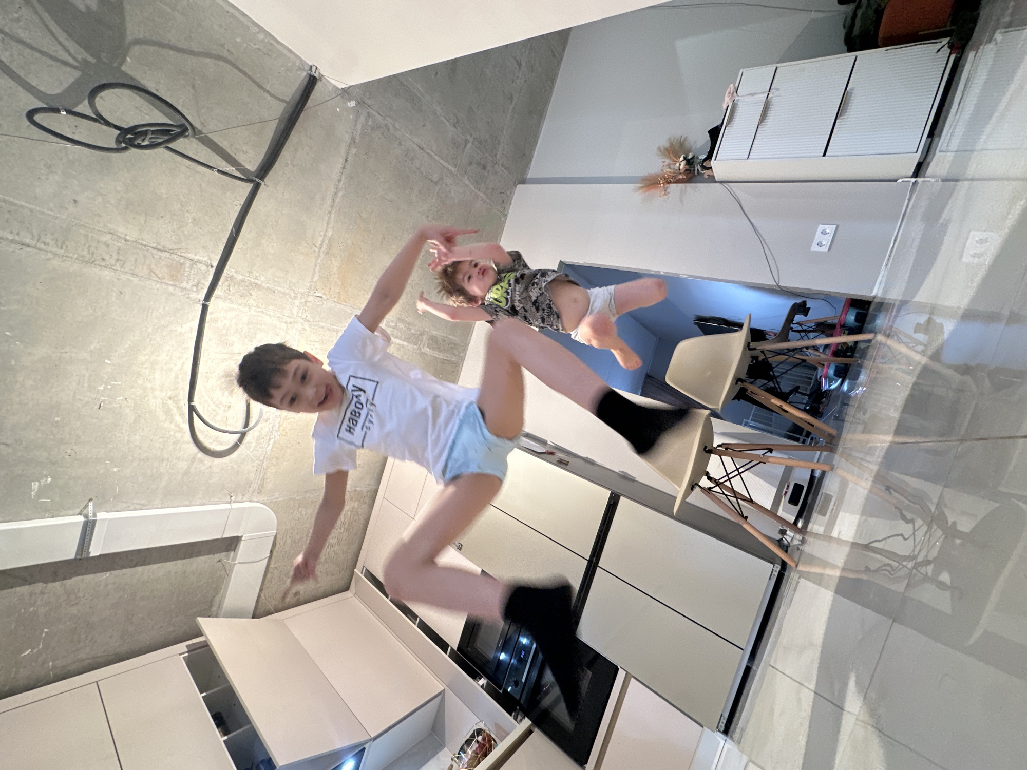 Two boys are captured mid-air in what appears to be a playful jump or levitation within a modern indoor setting. The image is significantly rotated clockwise, creating the illusion of the subjects floating horizontally.
The older boy, closer to the foreground, wears a white T-shirt, light blue shorts or underwear, and black socks/footwear. He is smiling broadly towards the camera, with his arms and legs dynamically extended. Visible text on his T-shirt appears to be in Cyrillic, reading "НАВСЕГДА" (meaning "forever" in Russian), which aligns with the provided Moscow, Russia context.
Behind him and slightly higher in the frame, a younger boy is also suspended mid-air. He wears a patterned T-shirt and a white diaper or underwear, with his limbs splayed out, engaged in the same playful activity. His expression is less clear but suggests concentration.
The background reveals a contemporary interior space, likely a kitchen or open-plan living area. Above the boys is a raw concrete ceiling with exposed black electrical conduits forming a decorative loop and a white ventilation duct running along it. Below them are sleek, light-colored kitchen cabinets, some with drawers open, and a built-in dark oven. A reflective tiled floor is visible, showing reflections of light-colored chairs with wooden legs. A white electrical outlet is mounted on a wall. The lighting is bright, illuminating the scene and casting shadows.