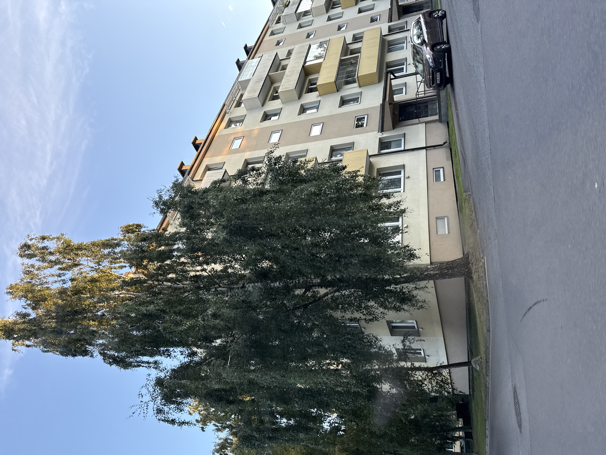 A tall, leafy tree with drooping branches is in the foreground, obscuring a portion of a multi-story apartment building. The building is light beige and tan, with several balconies, some of which are enclosed with glass. The building has numerous windows, some of which are illuminated. A dark-colored SUV is parked in front of the building. The scene appears to be a residential area in Tallinn, Estonia. The sky is clear and blue with wispy white clouds. The time of day is likely late afternoon or early evening, given the low angle of the sunlight casting warm tones on the building.