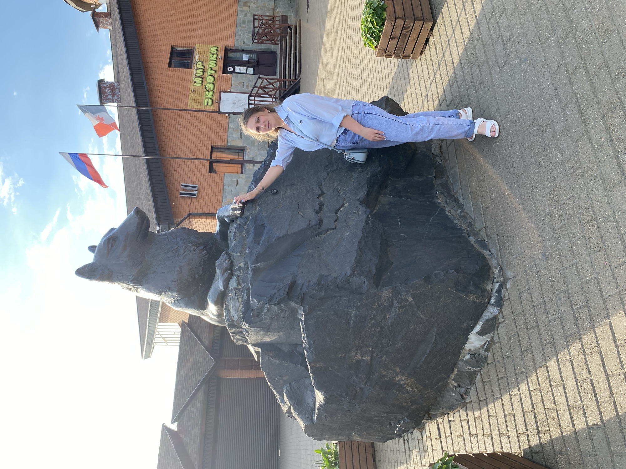 A young woman stands next to a large rock sculpture of a wolf in front of a brick building with a sign that reads "МИР ЭКЗОТИКИ" (World of Exotics) in Izhevsk, Russia. The woman is wearing a light blue button-up shirt and light blue jeans, with white sandals. She has a light blue purse slung over her shoulder. The wolf sculpture is dark gray and appears to be made of metal, sitting atop a rough, dark gray boulder. Two flags, the Russian flag and a red and white flag, fly above the building. The ground is paved with gray bricks, and there are wooden planters with green foliage to the left and right of the sculpture. The sky is blue with some white clouds, and the sun is casting shadows.