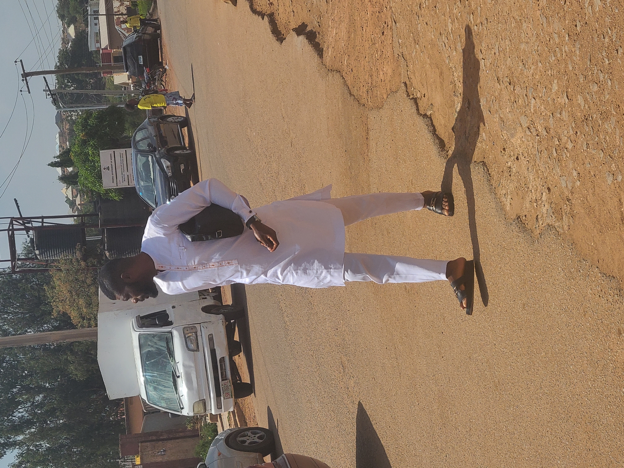 A person in a white outfit and black sandals is standing on a paved road. His shadow is cast to his right. In the background, there is a white truck, a grey car, and a black car, as well as street poles and buildings. There is a sign in the background that reads "FEDERAL GOVERNMENT OF NIGERIA".
