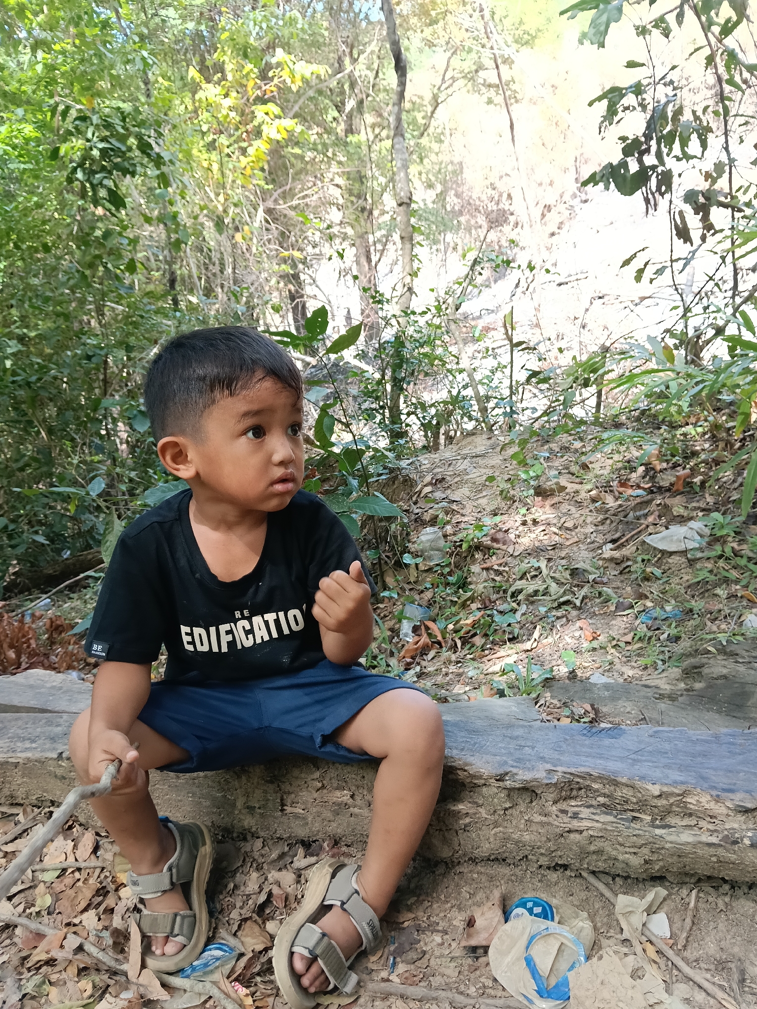 A young boy sits on a fallen log in a natural environment with dense foliage. He is wearing a black t-shirt and blue shorts, with sandals on his feet. The boy is holding a stick and looking to his right with a thoughtful expression. The background consists of trees, dried leaves, and some debris. The scene appears to be a rural or natural setting, potentially near a wooded area. The image does not provide specific information to determine the location in the city of Lampageu, Indonesia.
