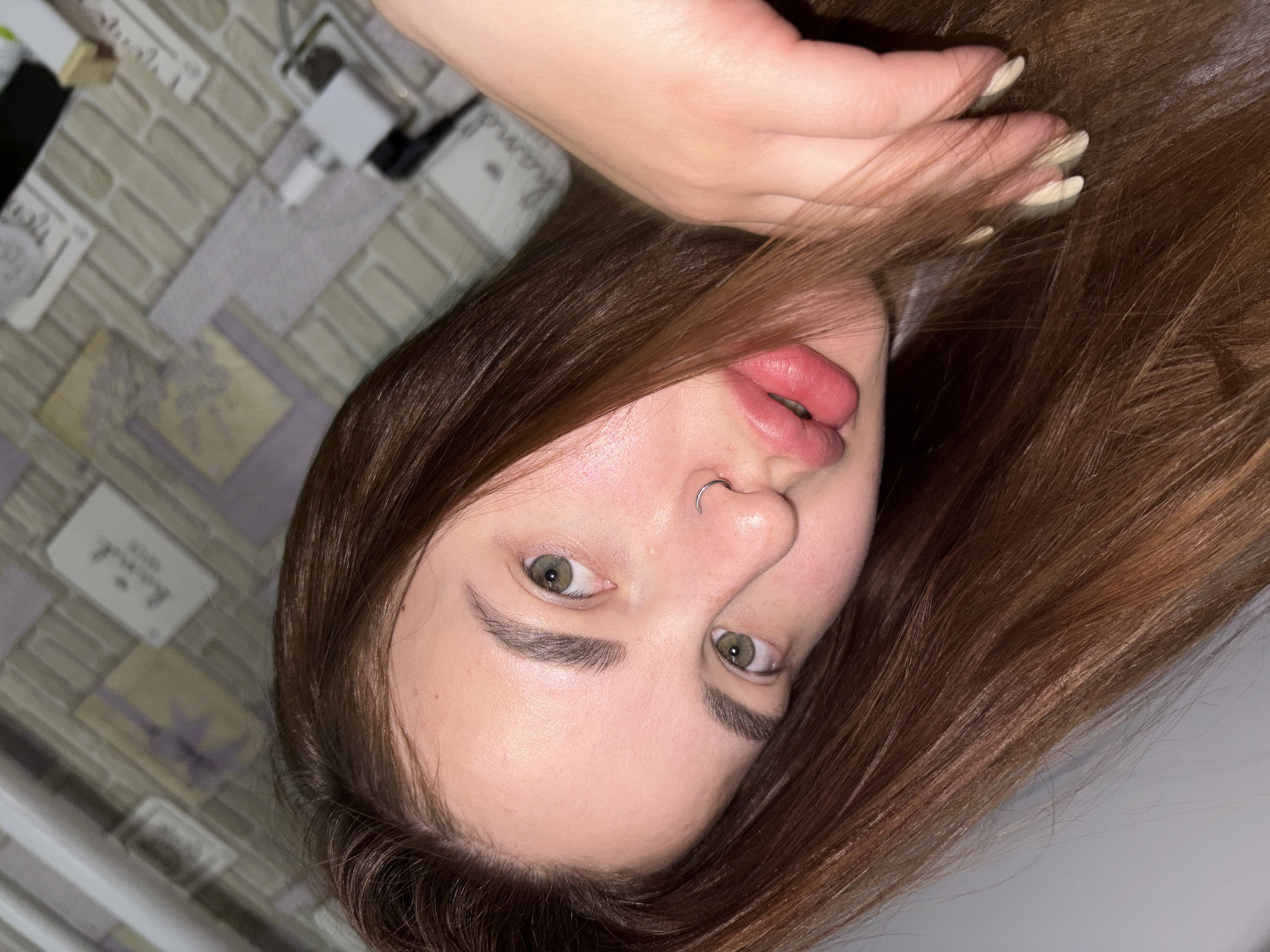 A selfie shows a young woman with light skin, green eyes, and brown hair. She has a nose ring in her left nostril. Her lips are full and pink. Her hair is styled and falls around her face and hand. The background is a blurred brick-like pattern with some items on it. The location, Port-Katon, Russia is not discernible from this image.