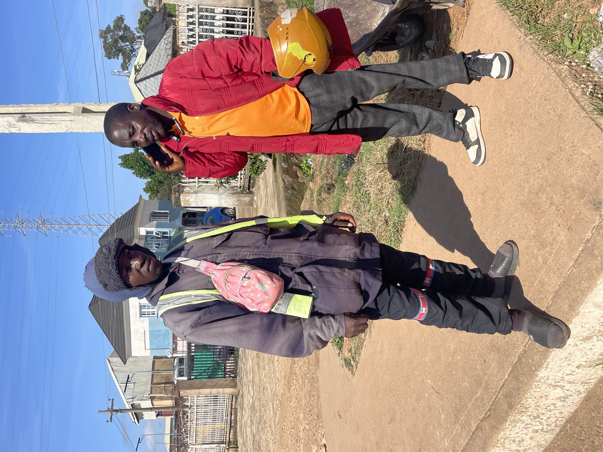 Two men are standing on a paved surface outdoors. The man on the left is wearing a dark blue hooded jacket with a fur lining, a reflective yellow vest over it, black pants with red and blue accents, and dark gray slide sandals. He has a small pink bag with a cartoon pattern slung across his chest. The man on the right is wearing a bright red jacket over an orange polo shirt and checkered gray pants. He is holding a yellow helmet in his right hand and talking on a dark cell phone with his left hand. They are in an urban environment with buildings, a power line tower, and fences visible in the background. The ground is a light brown, dusty earth. The lighting suggests it is daytime with shadows indicating sunlight.
