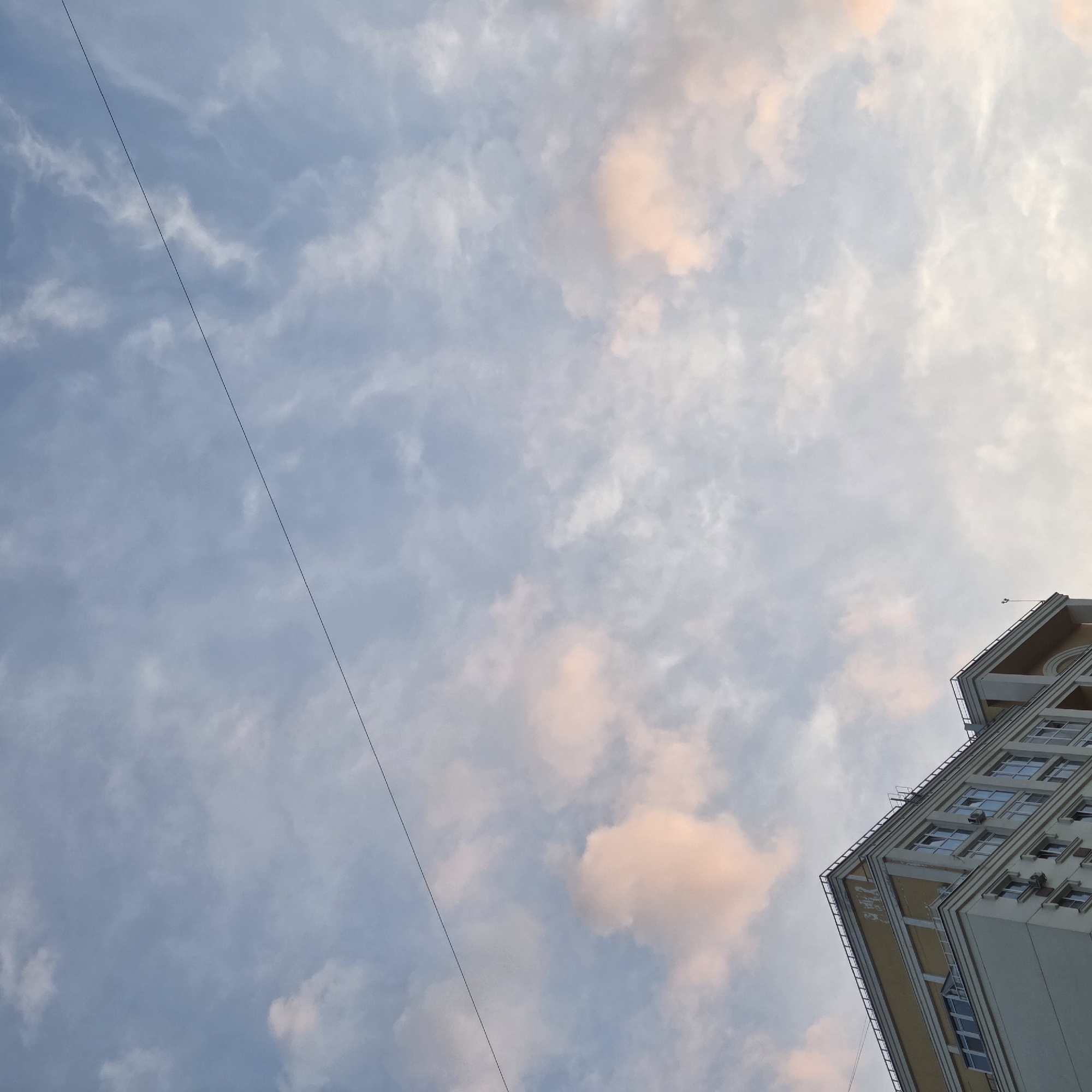 The image presents a low-angle view looking upwards at a partially cloudy sky and the corner of a building. The sky occupies the majority of the frame, displaying a palette of muted blue and soft grey tones. Scattered throughout are wispy clouds, many of which are illuminated with delicate pink and orange hues, strongly suggesting either a sunrise or a sunset.
A thin, dark wire or power line stretches diagonally across the sky from the upper left to the lower right of the frame.
In the bottom right corner, the upper part of a multi-story building is visible. The architecture appears modern or contemporary, with light-colored exterior walls, possibly off-white or light beige. Features include multiple rectangular windows, some reflecting the sky, and architectural details such as pronounced overhangs or cornices, the undersides of which are painted a contrasting golden or yellow-beige color. Small air conditioning units are mounted below some of the windows. On a vertical section of the building's facade, towards the right edge, there is some small, dark, indecipherable text or a logo.
The scene conveys a sense of calm, with the soft light of the sky contrasting with the structured lines of the urban environment. No people or specific activities are depicted.