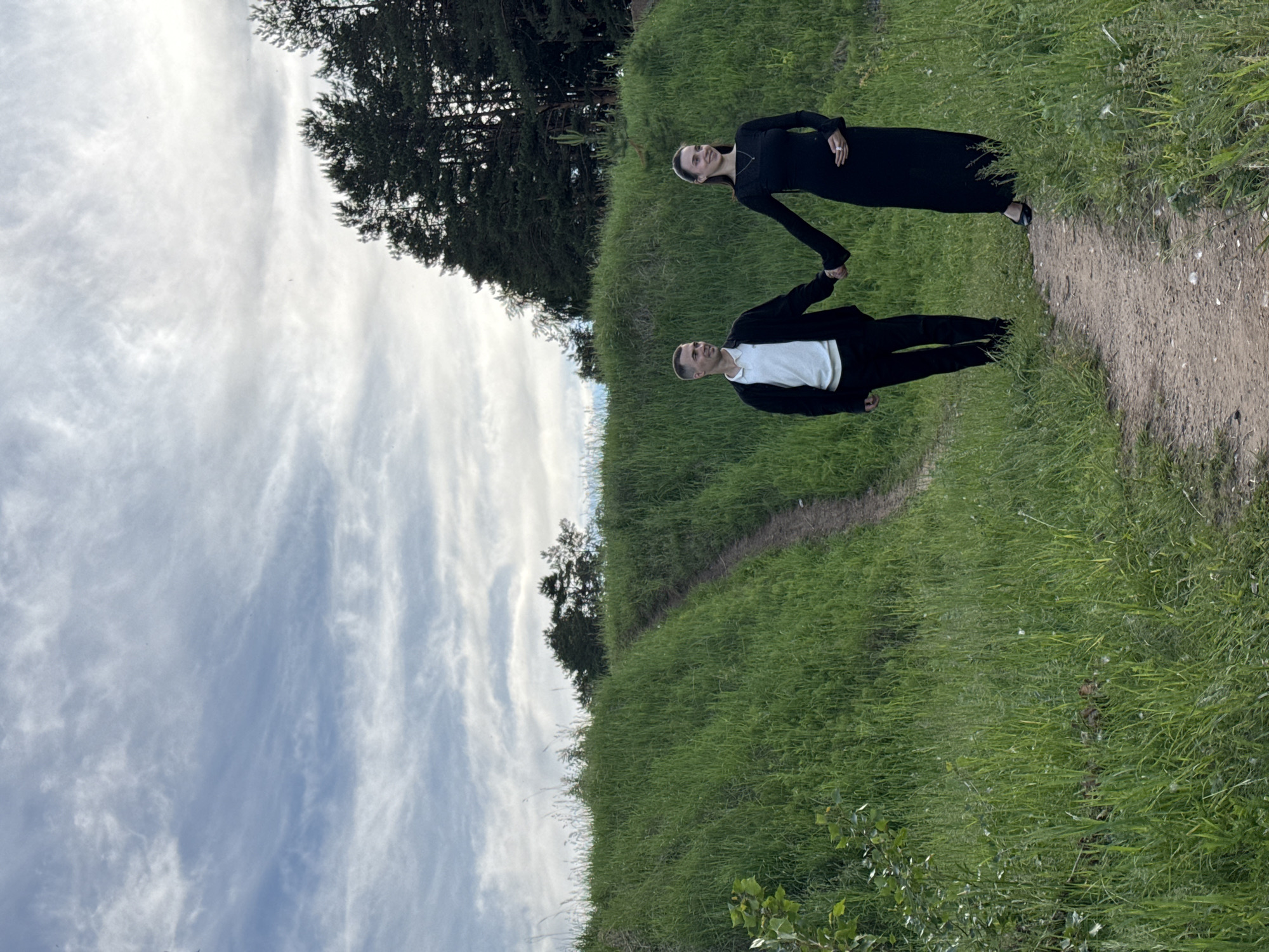The image captures a man and a woman standing hand-in-hand on a winding dirt path that cuts through a vibrant green grassy hillside. The scene is set outdoors under a cloudy sky, with a line of dark evergreen trees visible in the background.
The woman, positioned on the right side of the path, wears a long-sleeved black dress and black flat sandals. She has dark hair, likely pulled back, and is smiling broadly while looking towards the camera. A delicate necklace hangs around her neck, and a ring is visible on her right hand.
The man, on the left, is dressed in a white t-shirt layered under a black jacket, paired with black pants. He has short dark hair and is also smiling, looking in the same direction as the woman. Their clasped hands are central, emphasizing their connection.
The surrounding environment is characterized by dense green grass, indicating a fertile outdoor setting. The sky is filled with white, wispy clouds, suggesting an overcast or partly cloudy day, possibly in the late afternoon or early evening. The overall atmosphere appears pleasant and serene, with the couple exhibiting happiness and affection. No visible text is present in the image. The location context provided is Chaikovsky, Russia.