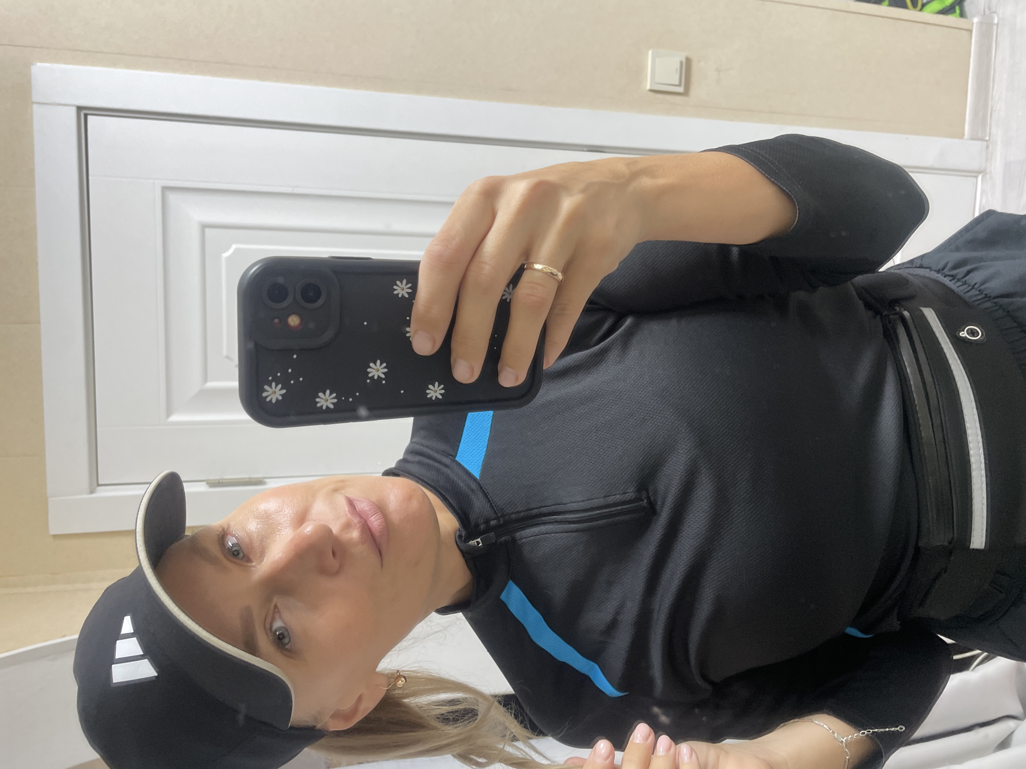 A woman with blonde hair and blue eyes is taking a selfie with a black smartphone in a black and white flowered phone case. She is wearing a black Adidas baseball cap with white stripes and a black and blue athletic top. A gold ring is visible on her left ring finger. She is standing in front of a white door with recessed panels and a white wall with a light switch. A silver anklet is visible on her left ankle. No information suggests the location of Baranovka, Russia.