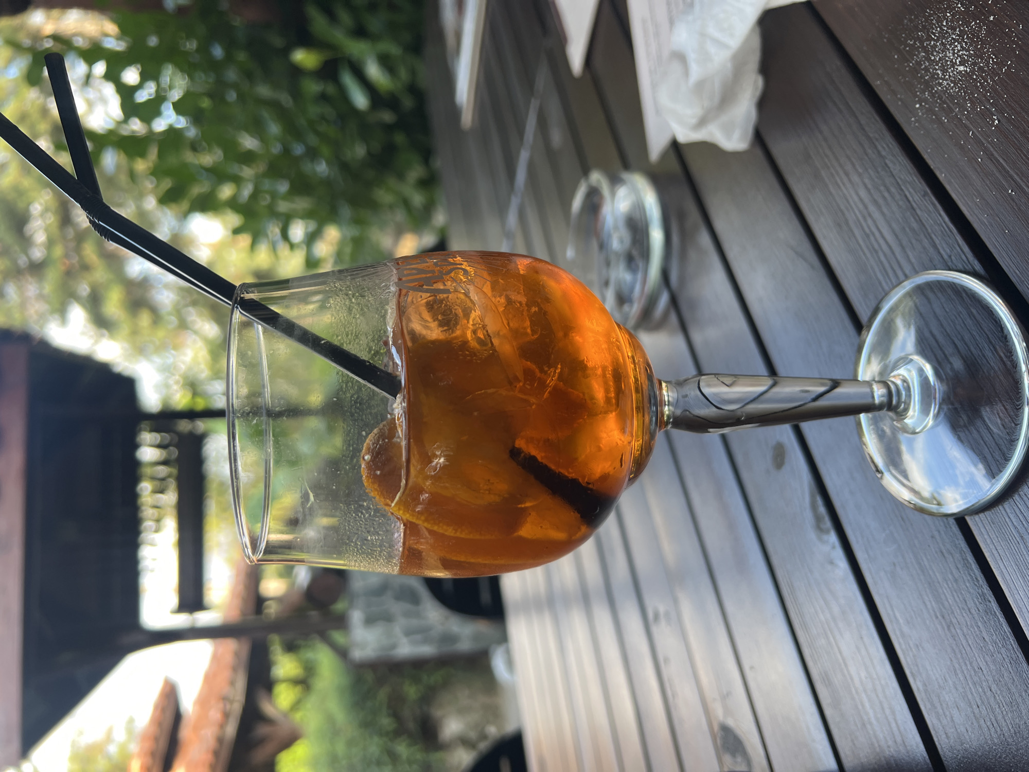 A close-up, eye-level shot captures an orange-colored alcoholic beverage in a wine glass, garnished with orange slices and ice, placed on a dark wooden table. Two black straws extend from the glass. In the background, out of focus, a covered outdoor seating area with a tiled roof is visible, surrounded by lush greenery. To the right of the glass, an ashtray and a crumpled white napkin rest on the table. The setting appears to be an outdoor dining establishment, possibly a restaurant or cafe, with hints of natural light filtering through the foliage. The overall ambiance suggests a relaxed outdoor setting in Plovdiv, Bulgaria, perhaps a terrace overlooking a garden or a picturesque courtyard. There are no people visible in the frame.