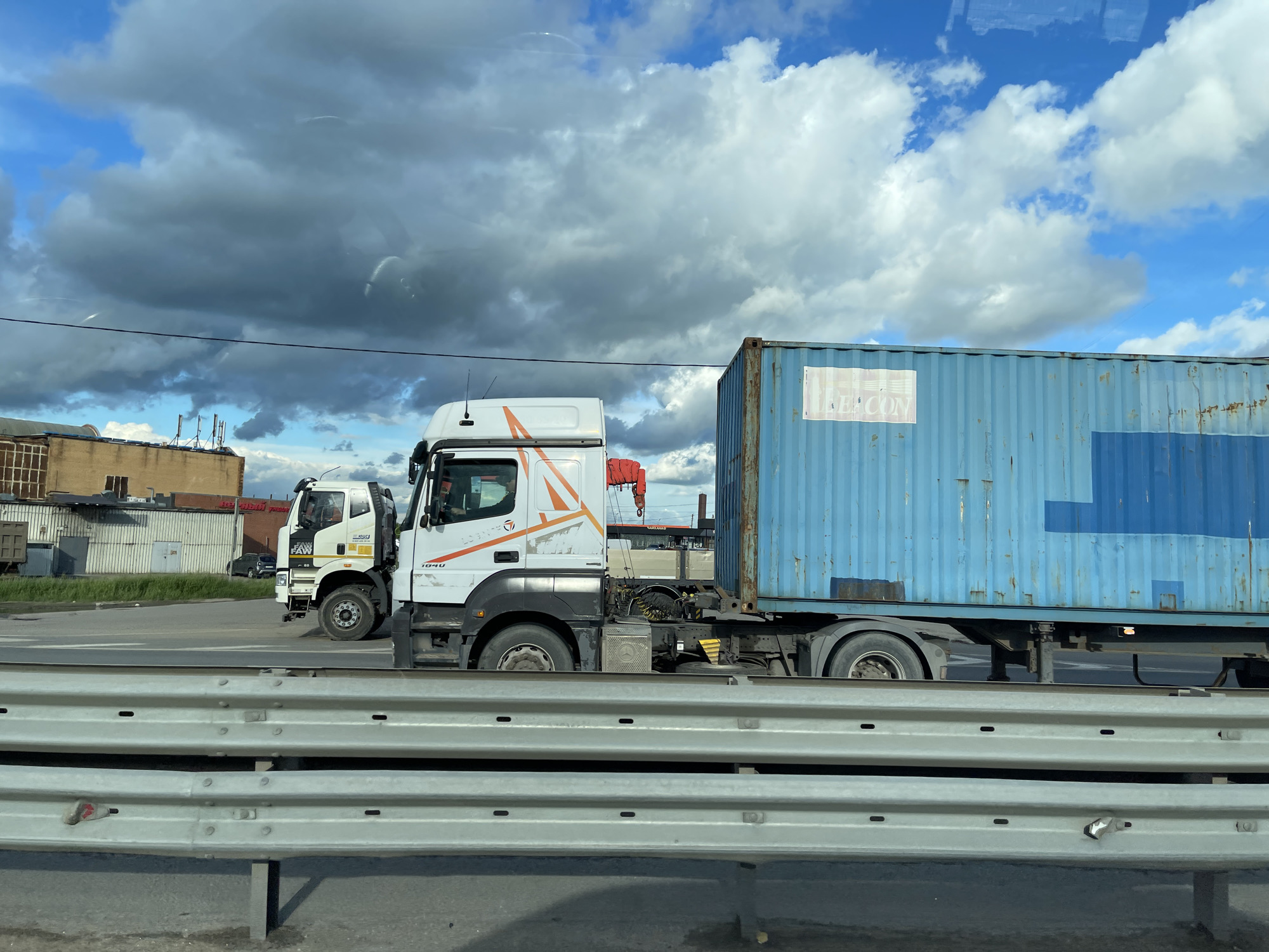 The image shows two white trucks on a road, with a blue shipping container attached to the closer truck. The truck has orange detailing and the text "L-TRANS 1040" on the side. Another truck is parked behind. The foreground shows a guard rail along the road. The background includes buildings and a cloudy sky with patches of blue. The image was taken in Podolsk, Russia.