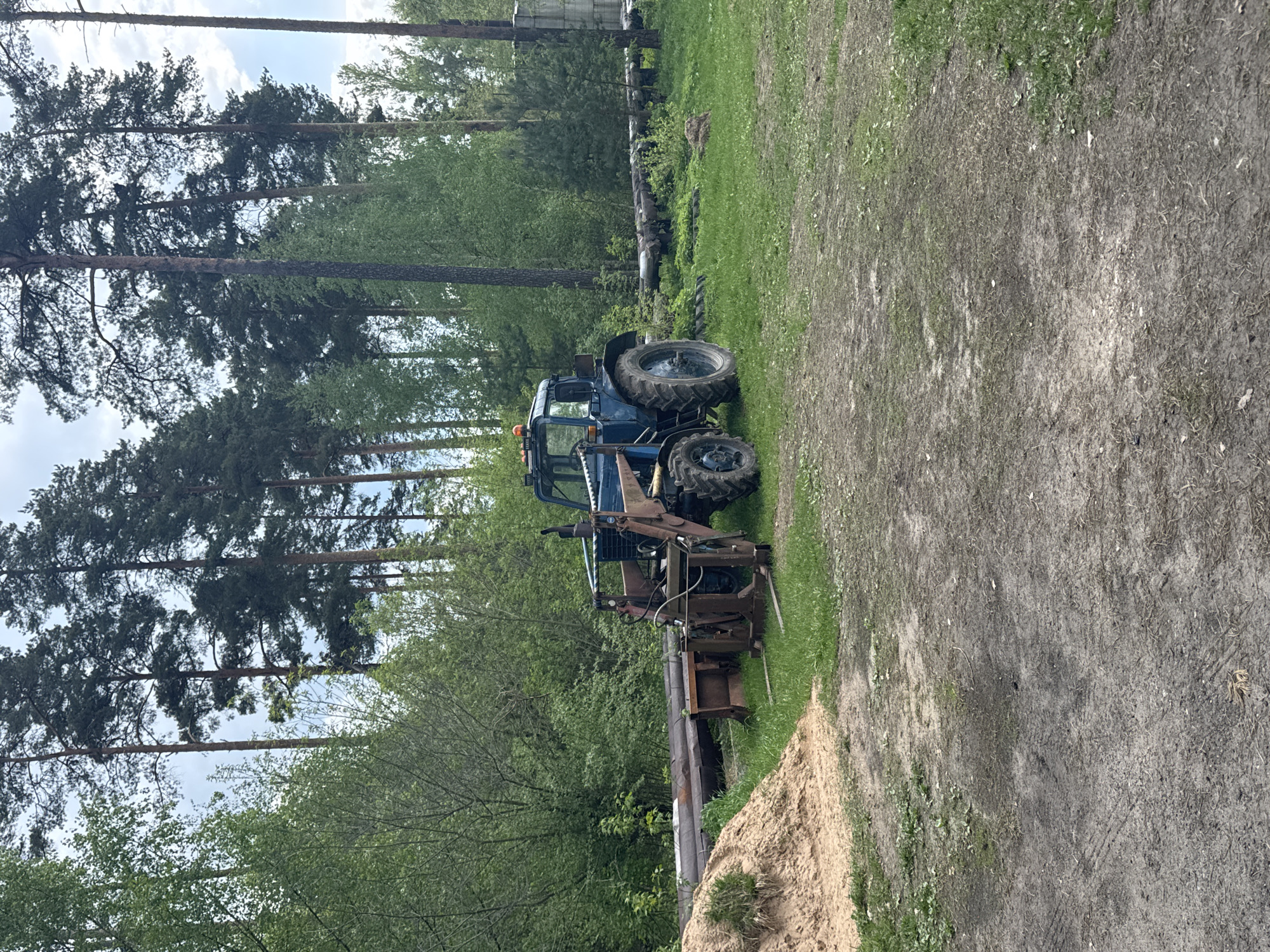 A blue tractor with an attached front-end loader is parked on a grassy area adjacent to a dirt path. The environment includes tall pine trees and a mix of other deciduous trees in the background, typical of a wooded area. There's a pile of sand or dirt visible near the tractor. The scene suggests a rural or semi-rural setting, given the presence of agricultural equipment and natural landscape. Based on the prompt and image analysis, the location is likely somewhere near Раздолье, Russia, which fits the described environment. No individuals are visible in the scene. The focus is on the tractor and its surroundings.