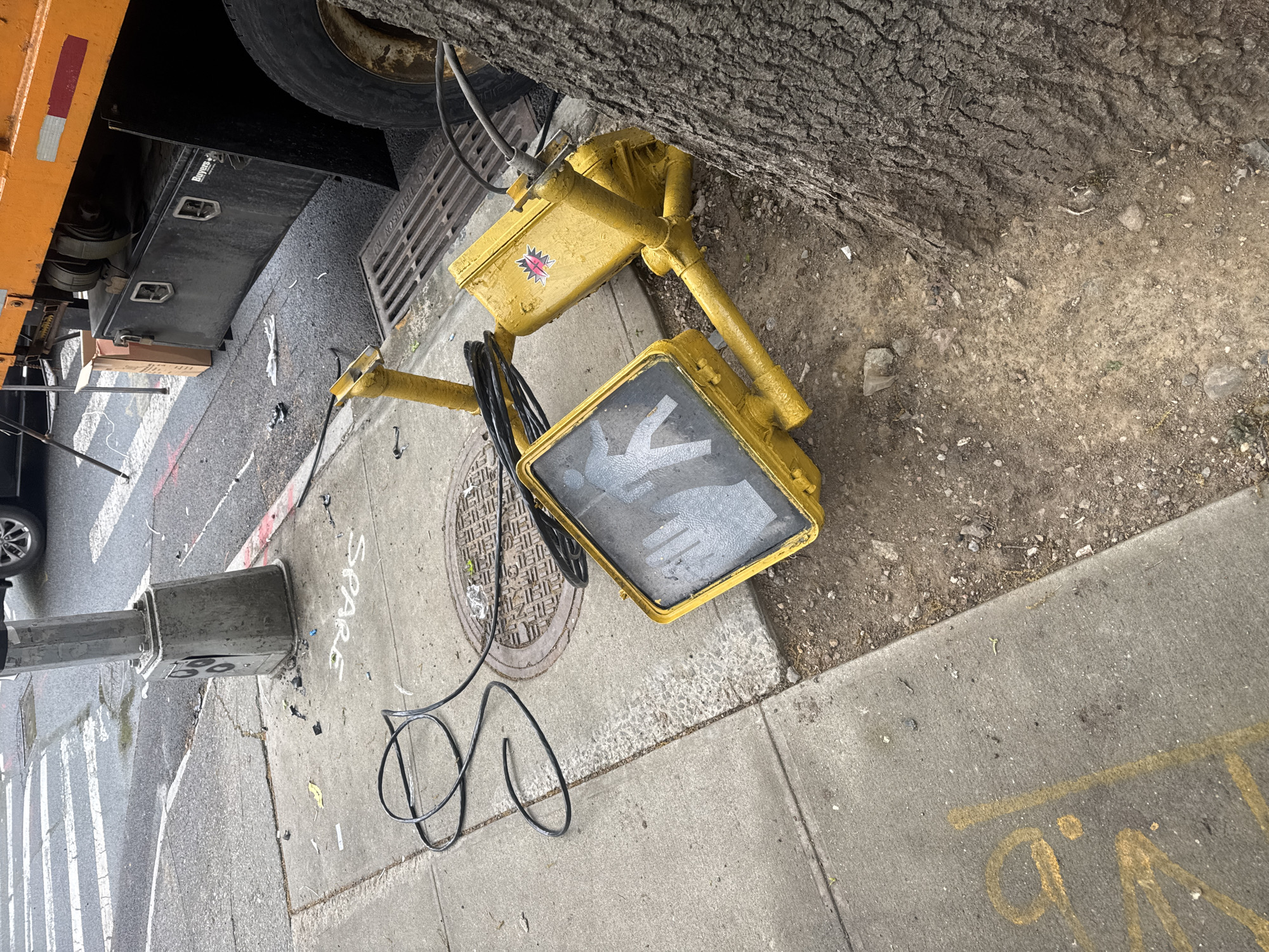 An outdoor, eye-level shot captures a dislodged pedestrian signal lying on the ground next to a large tree trunk, suggesting an urban incident or ongoing work in New York, USA. The bright yellow signal, featuring the "don't walk" (upraised hand) symbol, is dirty and its electrical wires are coiled on the concrete sidewalk. A red and white star-shaped sticker is affixed to the side of the signal box.
In the upper left, the undercarriage and a large tire of an orange vehicle, likely a truck, are visible. A black metal toolbox with the brand "Buyers" stamped on it is mounted beneath the truck. Next to the truck, a metal grate is embedded in the ground.
The scene includes a concrete sidewalk and an asphalt street. The sidewalk shows cracks and dirt, with white chalk or paint spelling "SPARE" near the base of a utility pole. Additional yellow markings, possibly "Q.D." or "O.D.", are on the sidewalk. Part of a manhole cover is visible, with the partial text "HARRISON NJ" readable, indicating its origin. The street features painted white crosswalk stripes and red and white lines, possibly temporary road markings. The ground around the tree is bare earth mixed with small stones and debris. The lighting suggests it is daytime, possibly overcast.